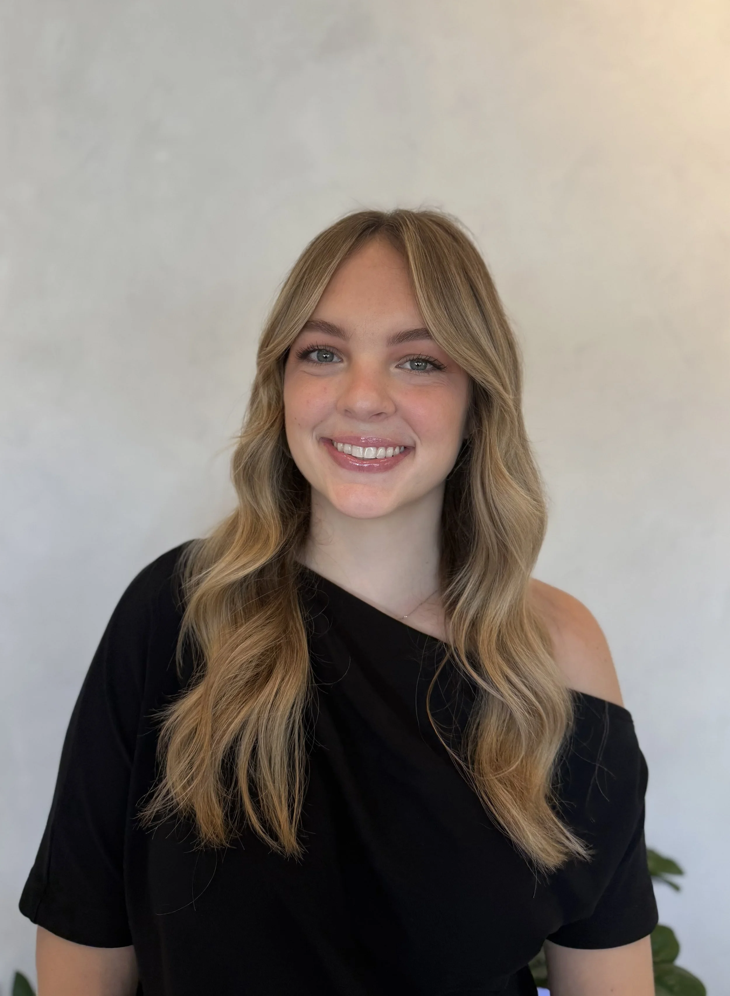 A young woman with long, wavy blonde hair smiling at the camera, wearing a black top with a cold shoulder detail, standing against a plain light-colored wall.