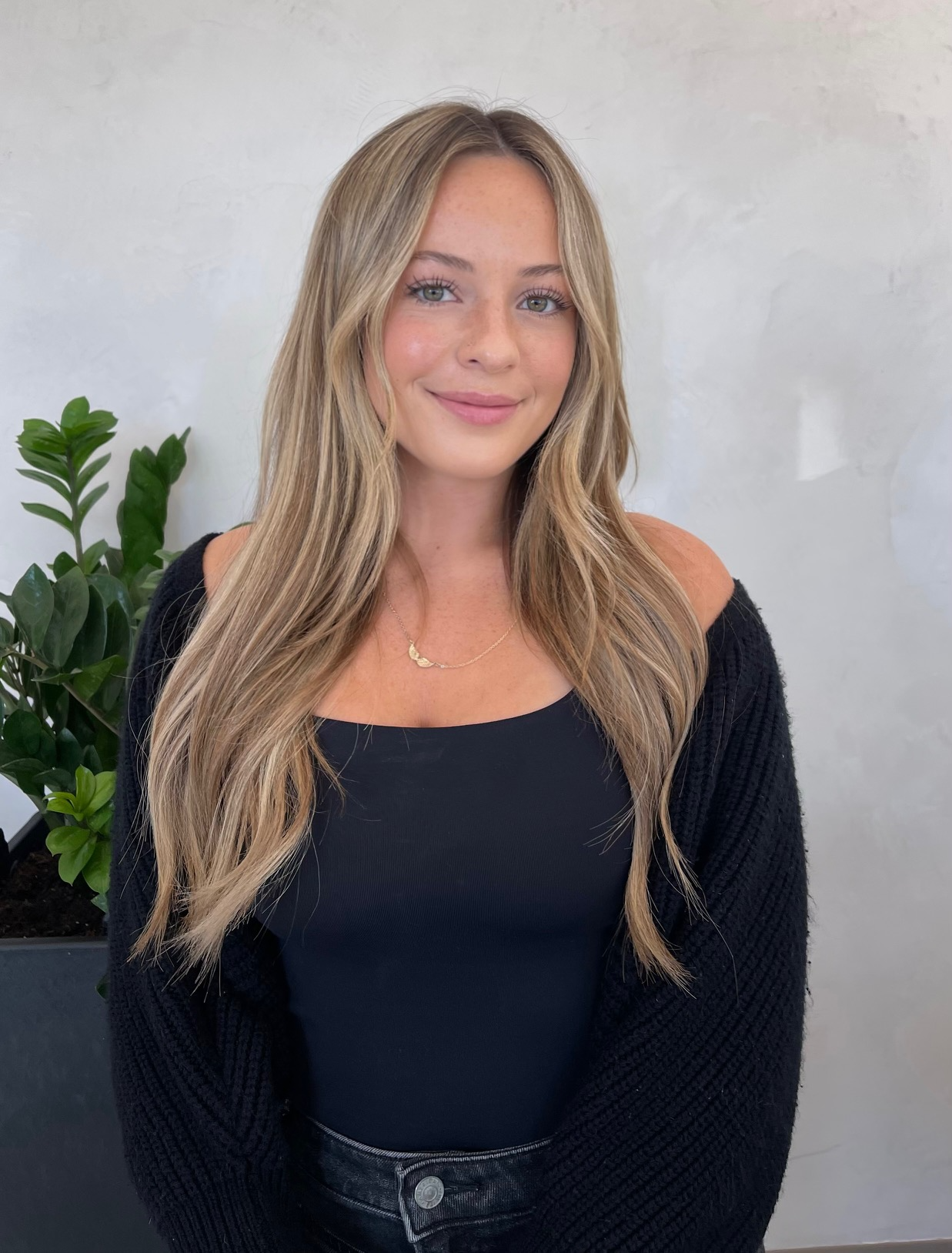 A young woman with long, wavy blonde hair, wearing a black top, a black cardigan, and a gold necklace, standing in front of a white wall and a green plant.