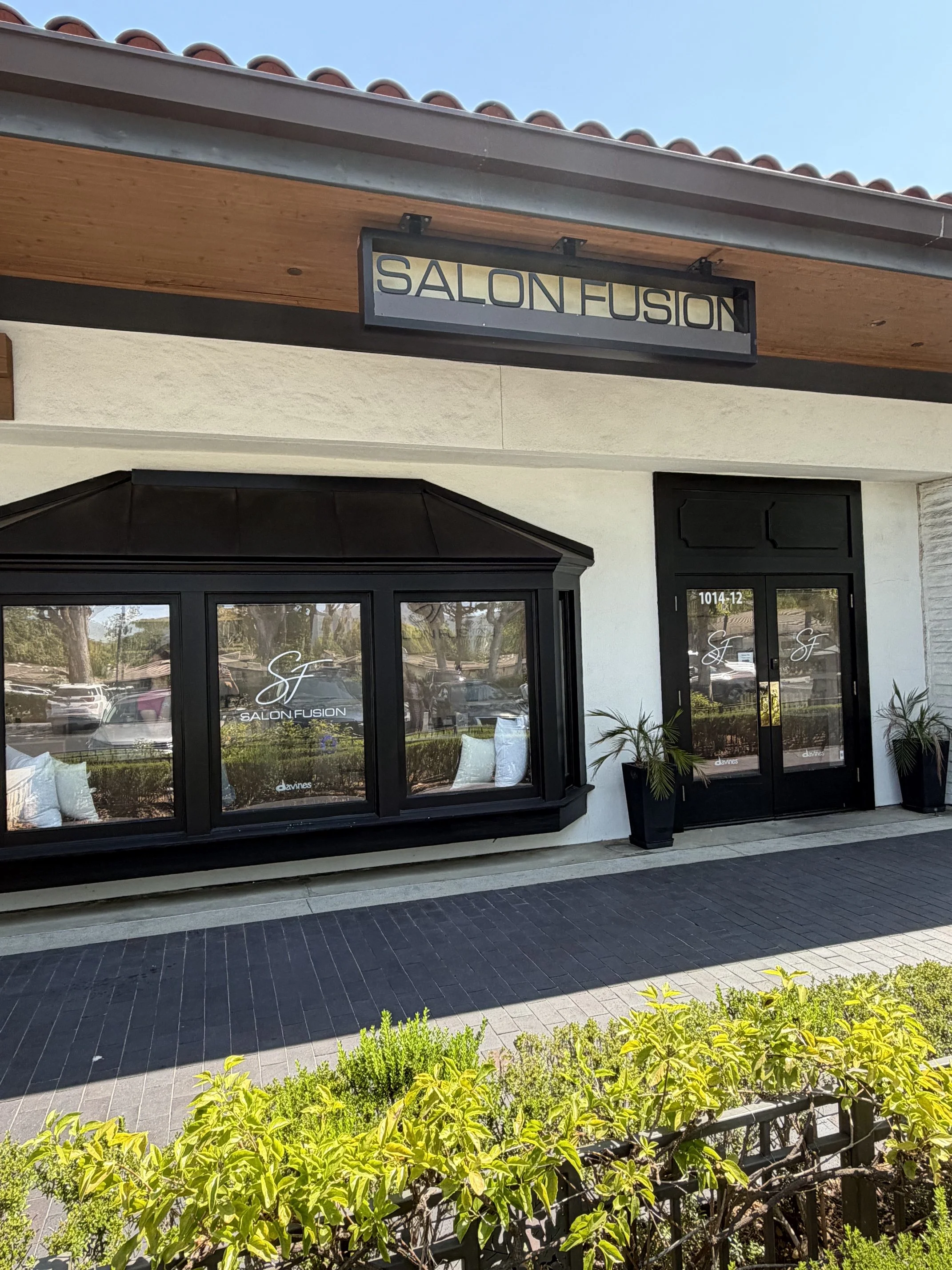 Exterior view of a storefront with black-framed glass doors and windows, labeled 'SALON FUSION,' with potted plants on either side and a landscaped foreground with green bushes and yellow flowers.