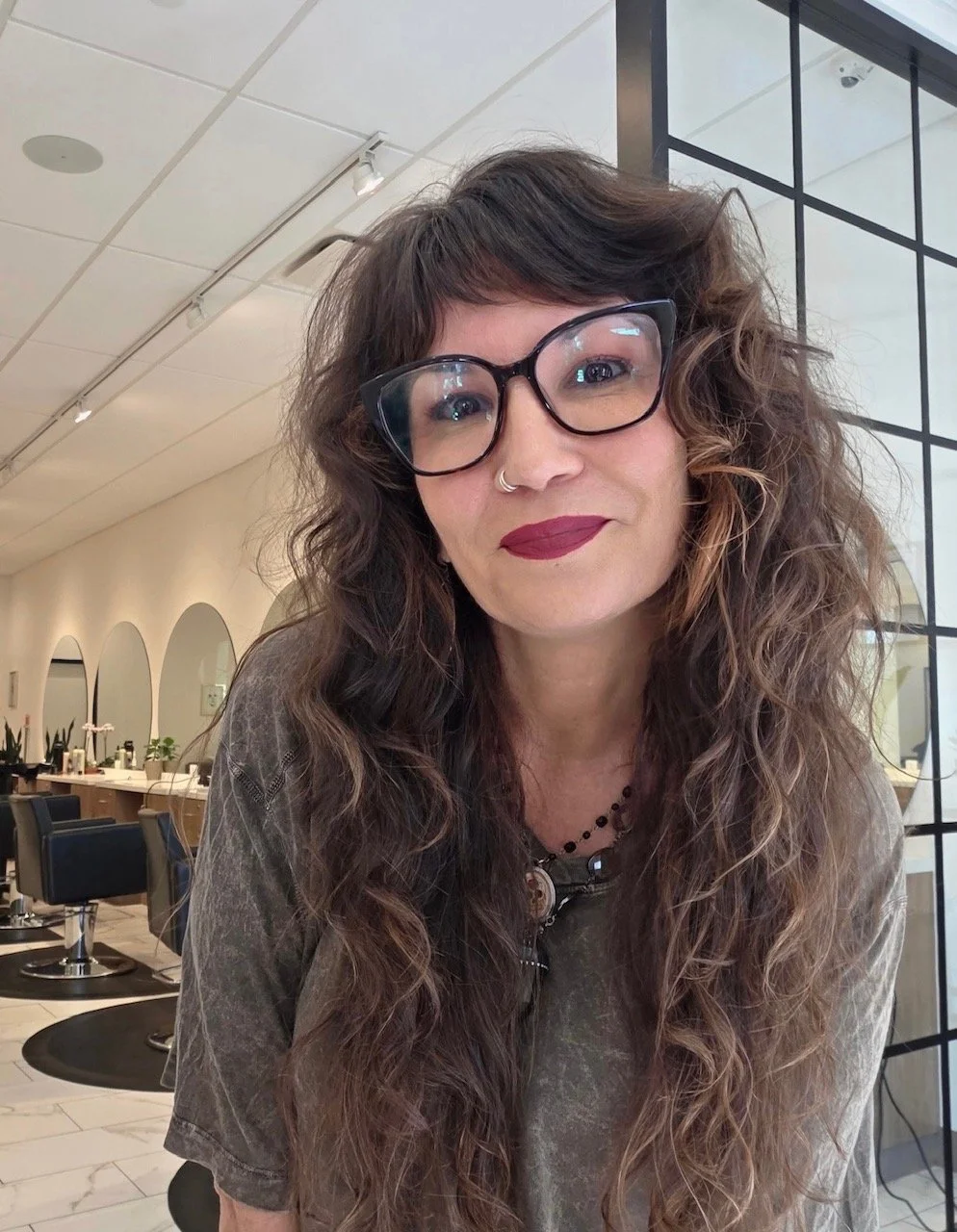 A woman with curly brown hair, glasses, and a nose ring, smiling in a salon or similar setting with black chairs and large mirrors in the background.