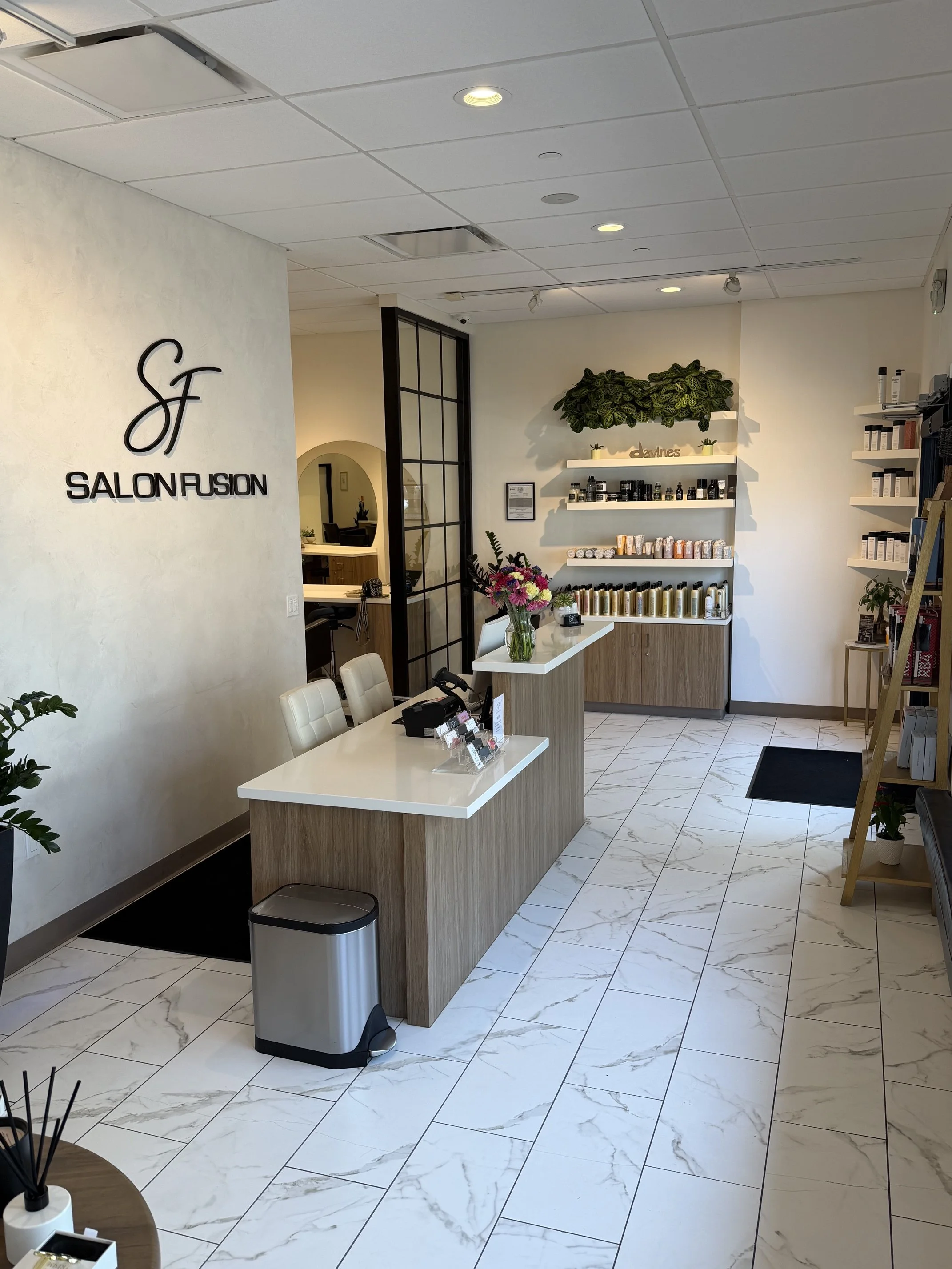 Interior of a beauty salon with a reception desk, shelves of products, a bouquet of flowers, and modern decor including white marble flooring and green plants.