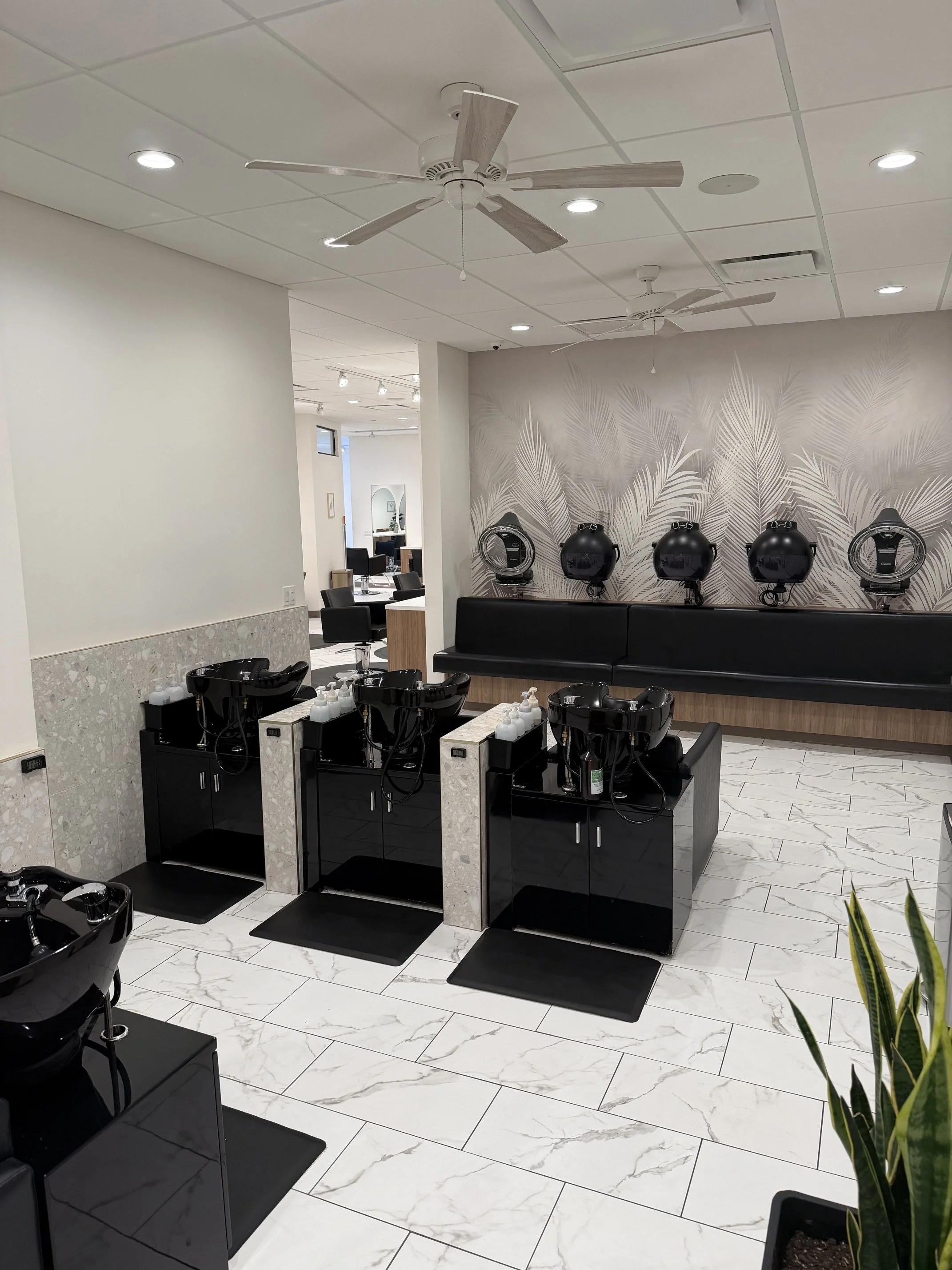 Interior of a modern hair salon with white marble flooring, black shampoo stations, and a beige wall with palm leaf design. Ceiling has white tiles and two ceiling fans, and there are various lights and mirrors visible in the background.