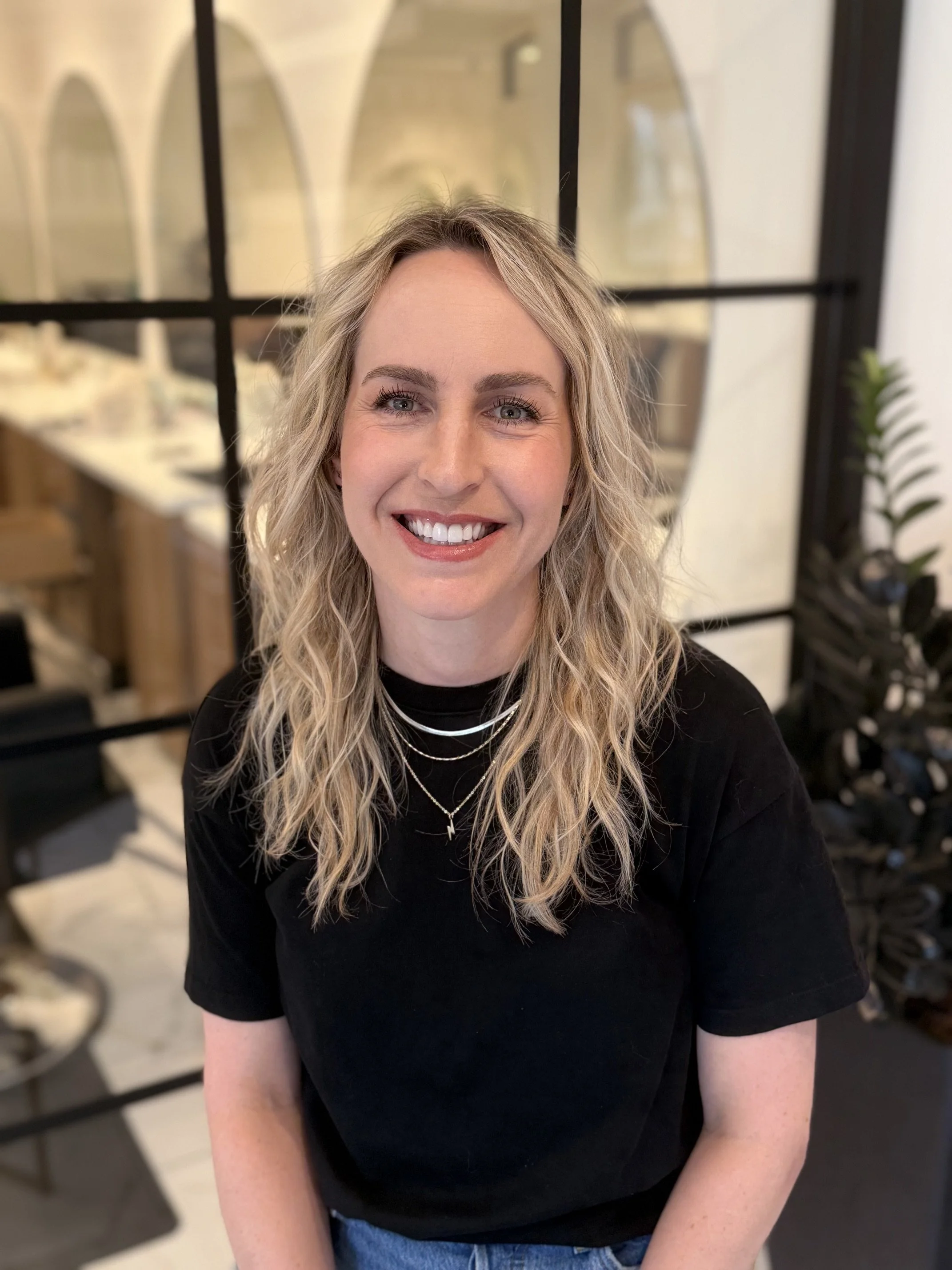 A smiling woman with blonde wavy hair wearing a black shirt and layered necklaces, sitting in a room with decorative arches and plants.