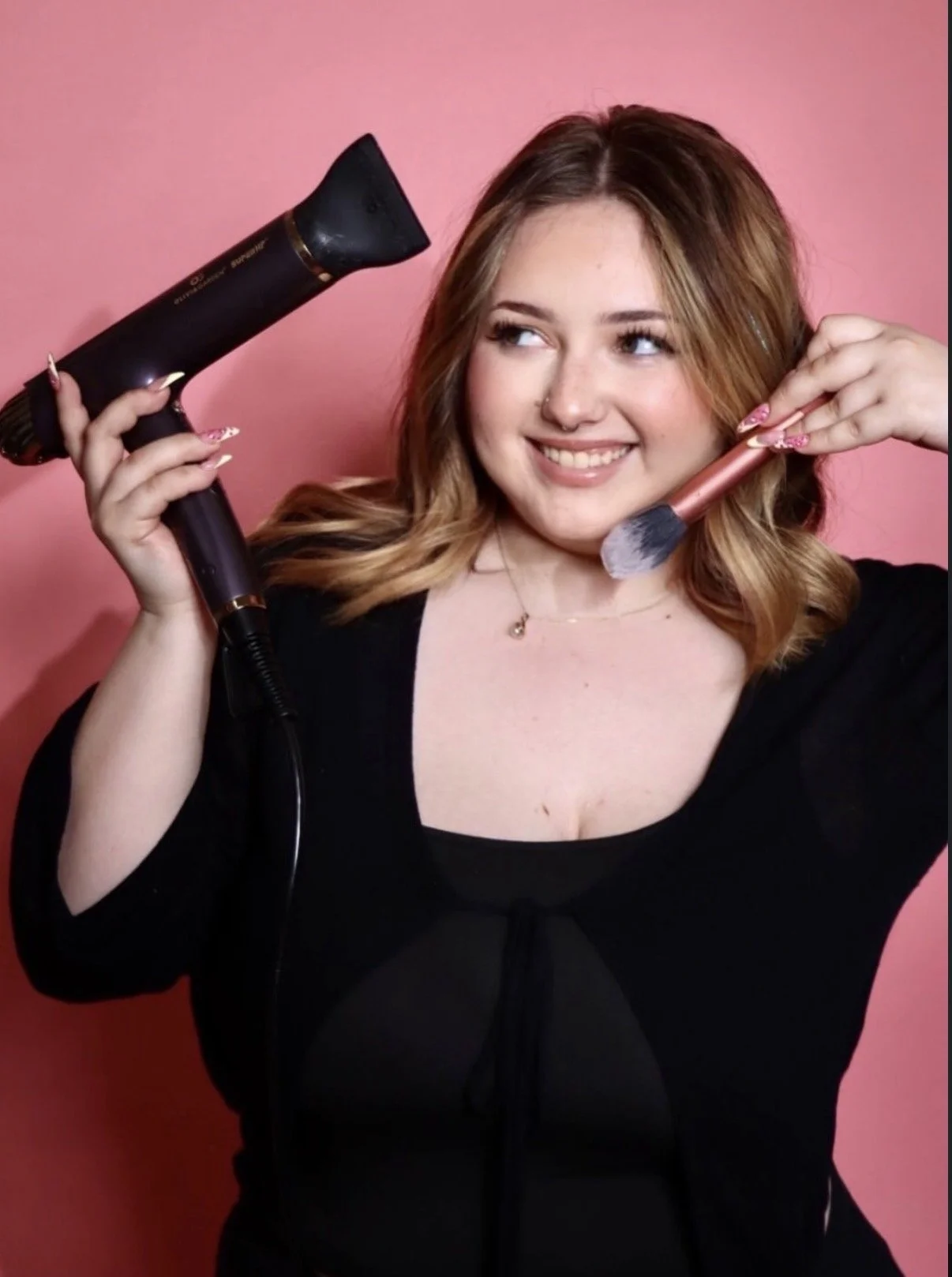 A smiling young woman with light brown, wavy hair holding a hairdryer in her right hand and a makeup brush in her left hand against a pink background.