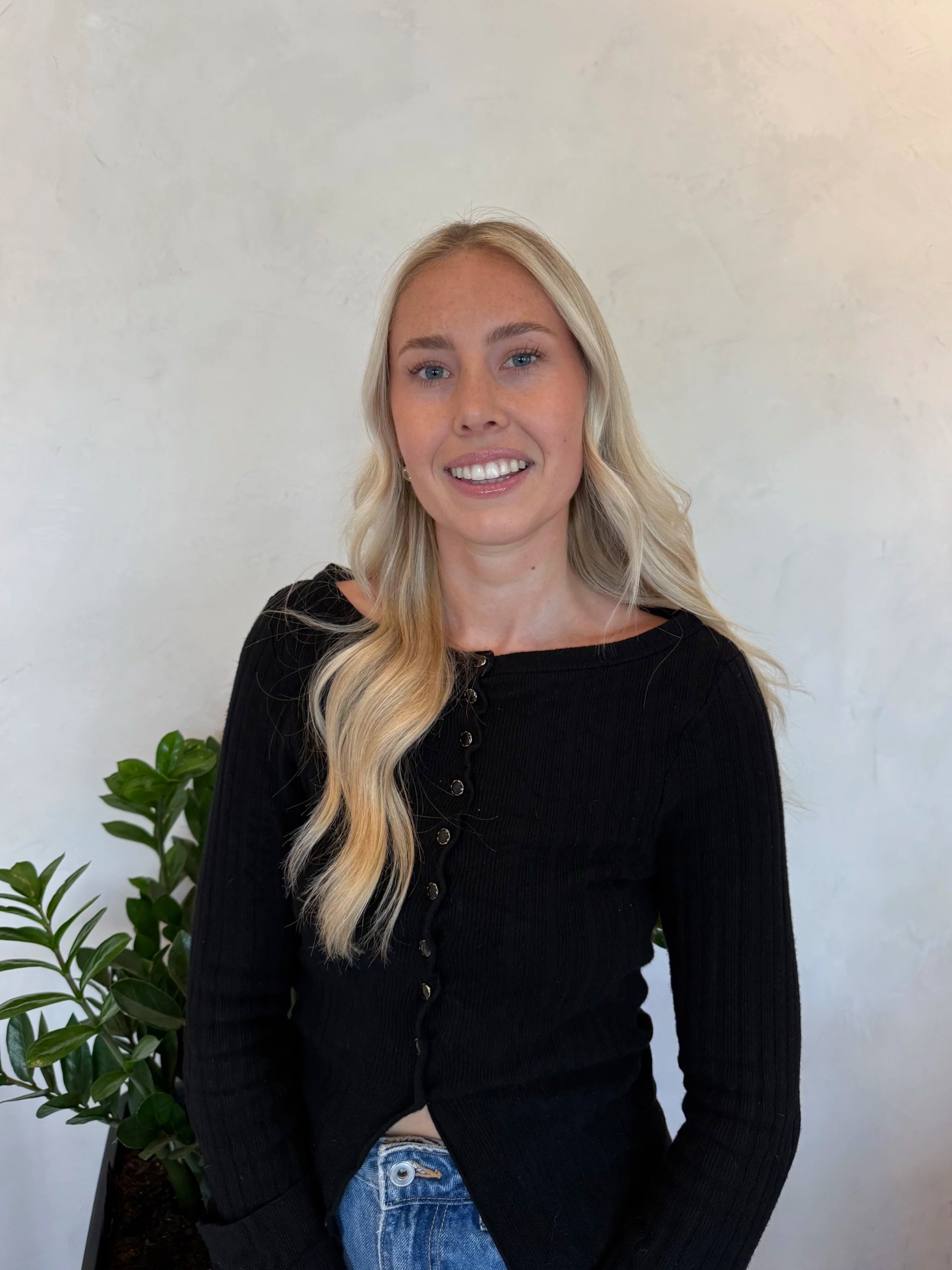 A woman with long blonde hair, blue eyes, and fair skin smiling, wearing a black button-up shirt and blue jeans, standing in front of a white wall with a green plant on her left.