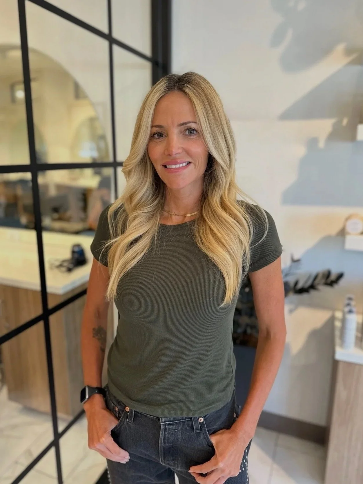 A smiling woman with long blonde hair, wearing a fitted olive green t-shirt, standing in a modern office space with glass partition and a desk in the background.