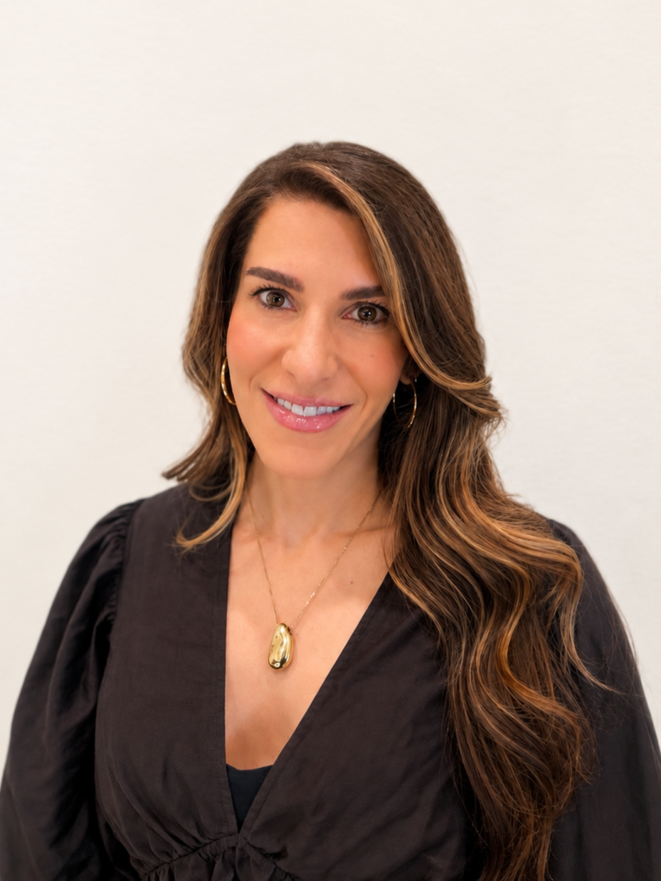 A woman with long, wavy brown hair is smiling at the camera, wearing a black top and gold jewelry, standing against a plain white wall.