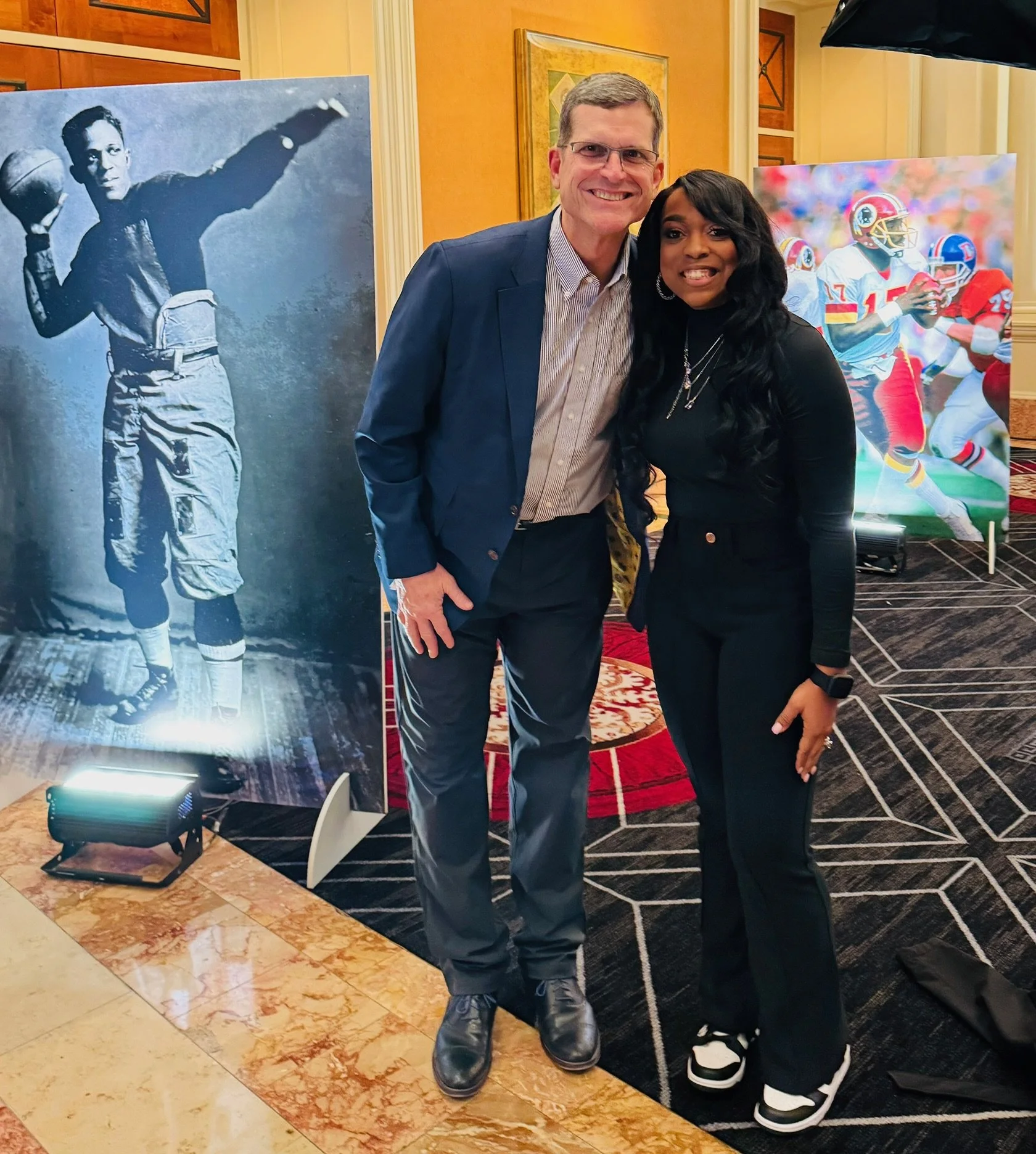 Two people, a man in a gray suit and a woman in all black, standing together and smiling at the camera in front of a display of sports posters, including a black-and-white photo of a football player and a colorful action shot of football players during a game.