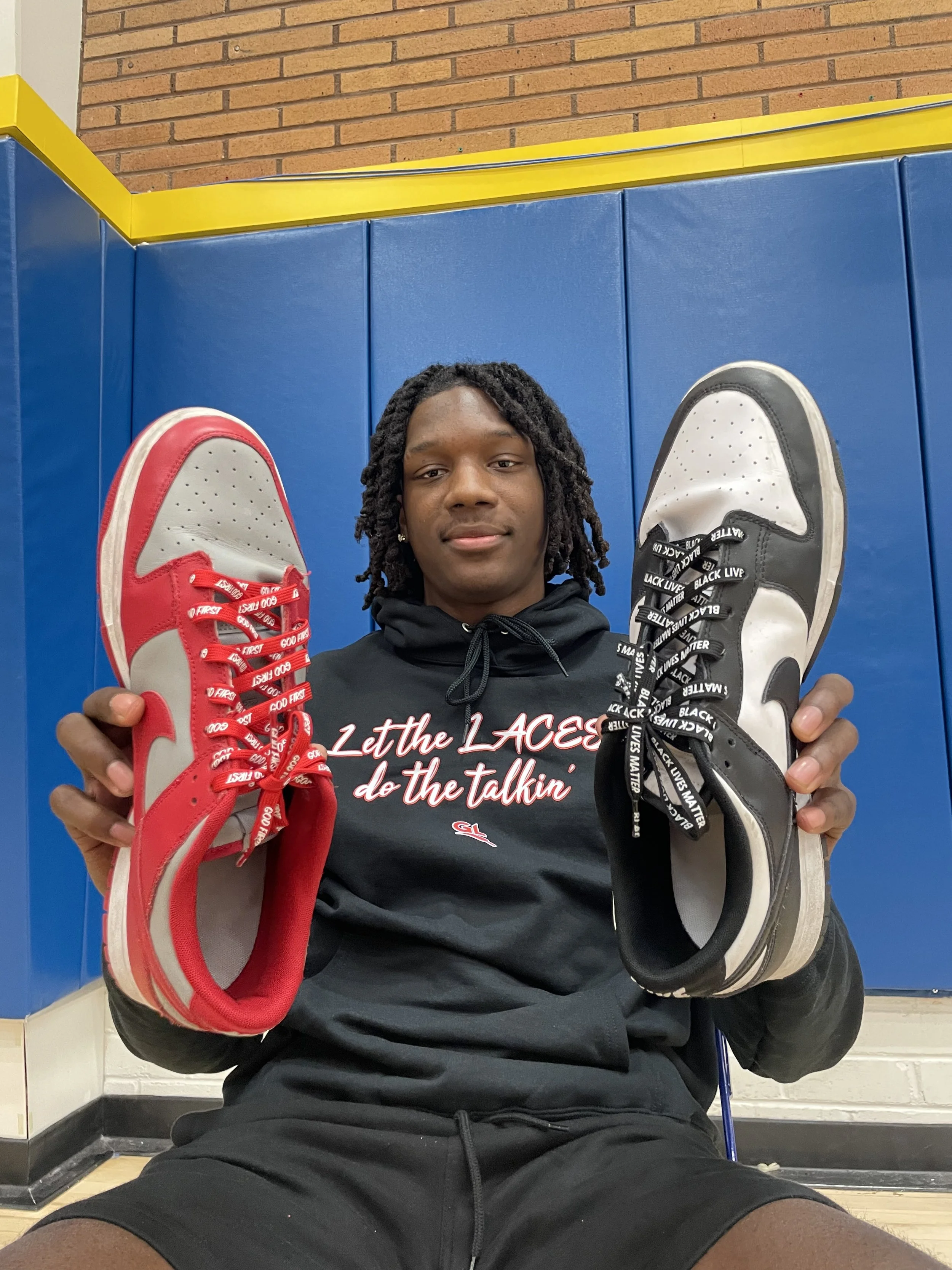 Young man with dreadlocks holding a red and a black and white sneaker, sitting in a gymnasium with blue padding and a brick wall behind him.