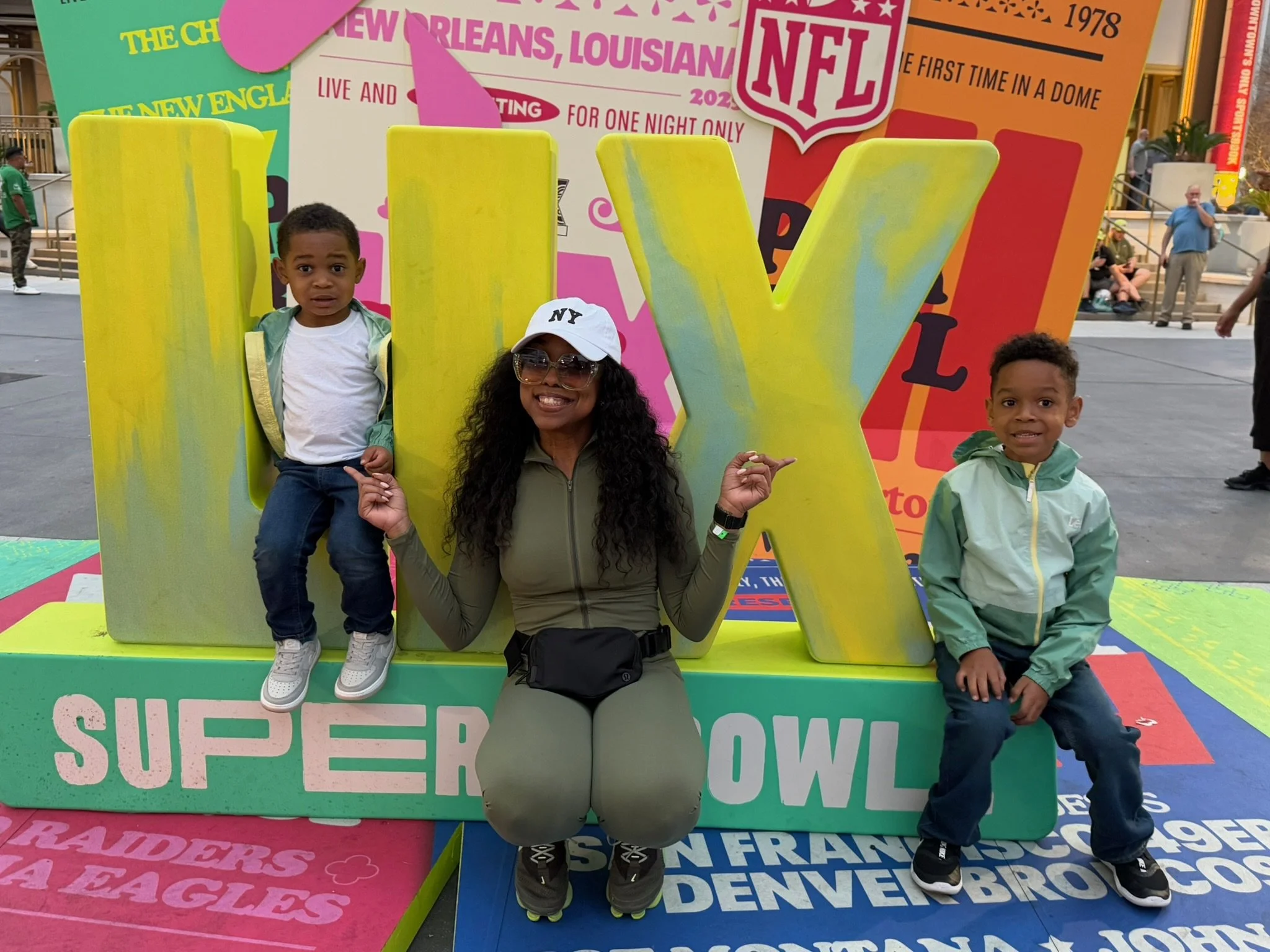 A woman and two children posing in front of large yellow letters spelling 'LII' at a Super Bowl event. The woman is kneeling, smiling, wearing sunglasses, a white cap, and a green jacket. The child on the left is sitting inside the left letter 'L', wearing a white shirt, green jacket, and jeans. The child on the right is sitting on the stage, wearing a green jacket and dark pants, smiling at the camera. The background features colorful posters and banners related to the Super Bowl and NFL.