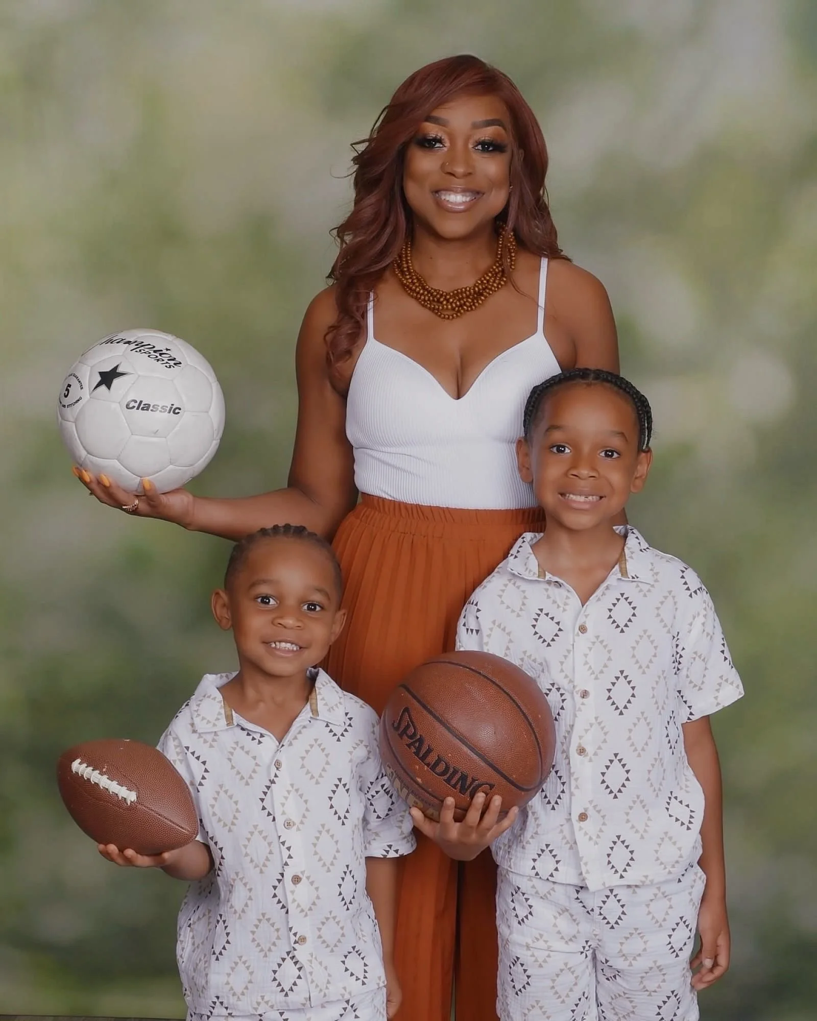 A woman with reddish-brown hair smiling, holding a white soccer ball in her right hand, standing with two young boys in matching patterned outfits, each holding a football, in front of a blurred outdoor background.