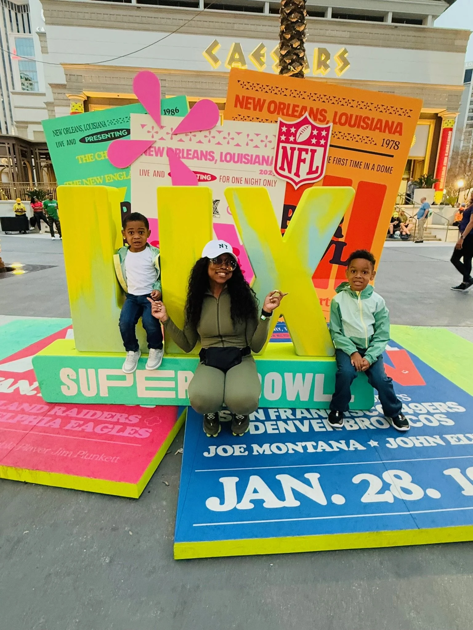 A woman and two children posing in front of colorful oversized NFL Super Bowl signs with tickets and event details displayed on the signs in Las Vegas.