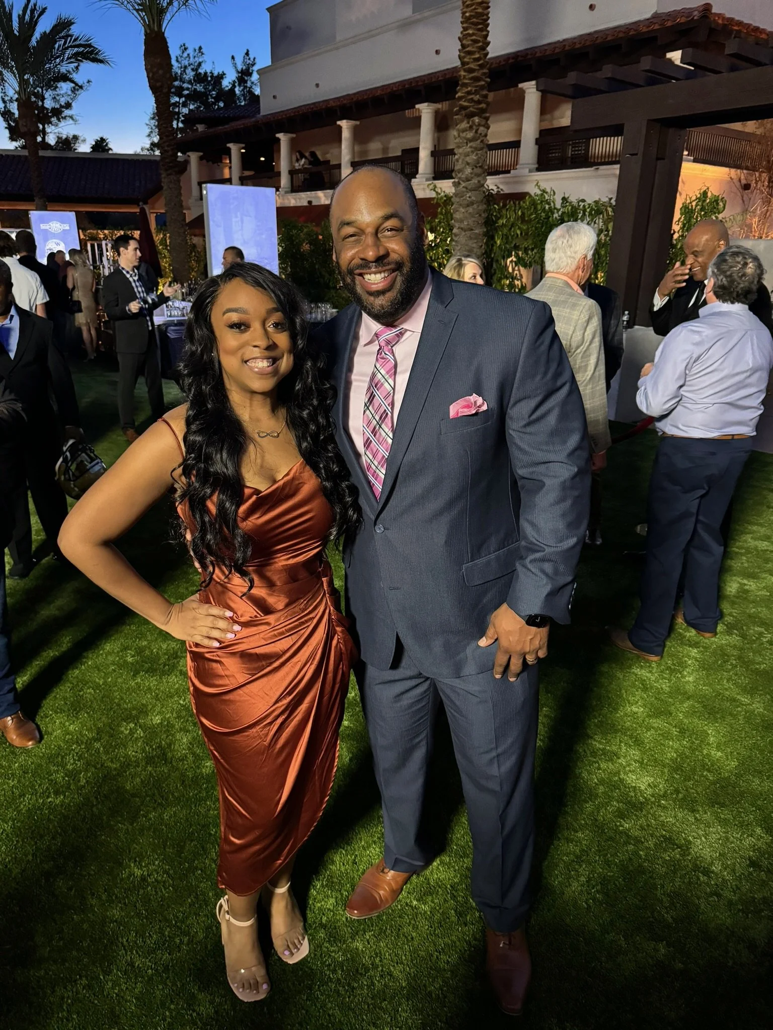 A woman in a satin copper-colored dress and a man in a gray suit with a pink tie, posing together at an outdoor evening event with other attendees in the background.