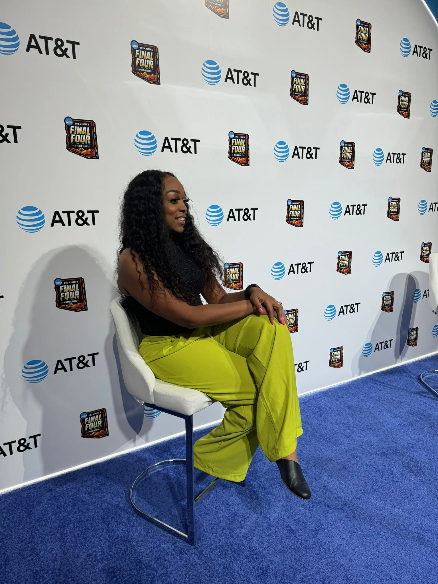 A woman with long curly hair, wearing a black sleeveless top and bright yellow pants, sitting on a white chair at an event with a backdrop featuring the AT&T logo and 'Final Four' logos for the 2024 men's basketball tournament.