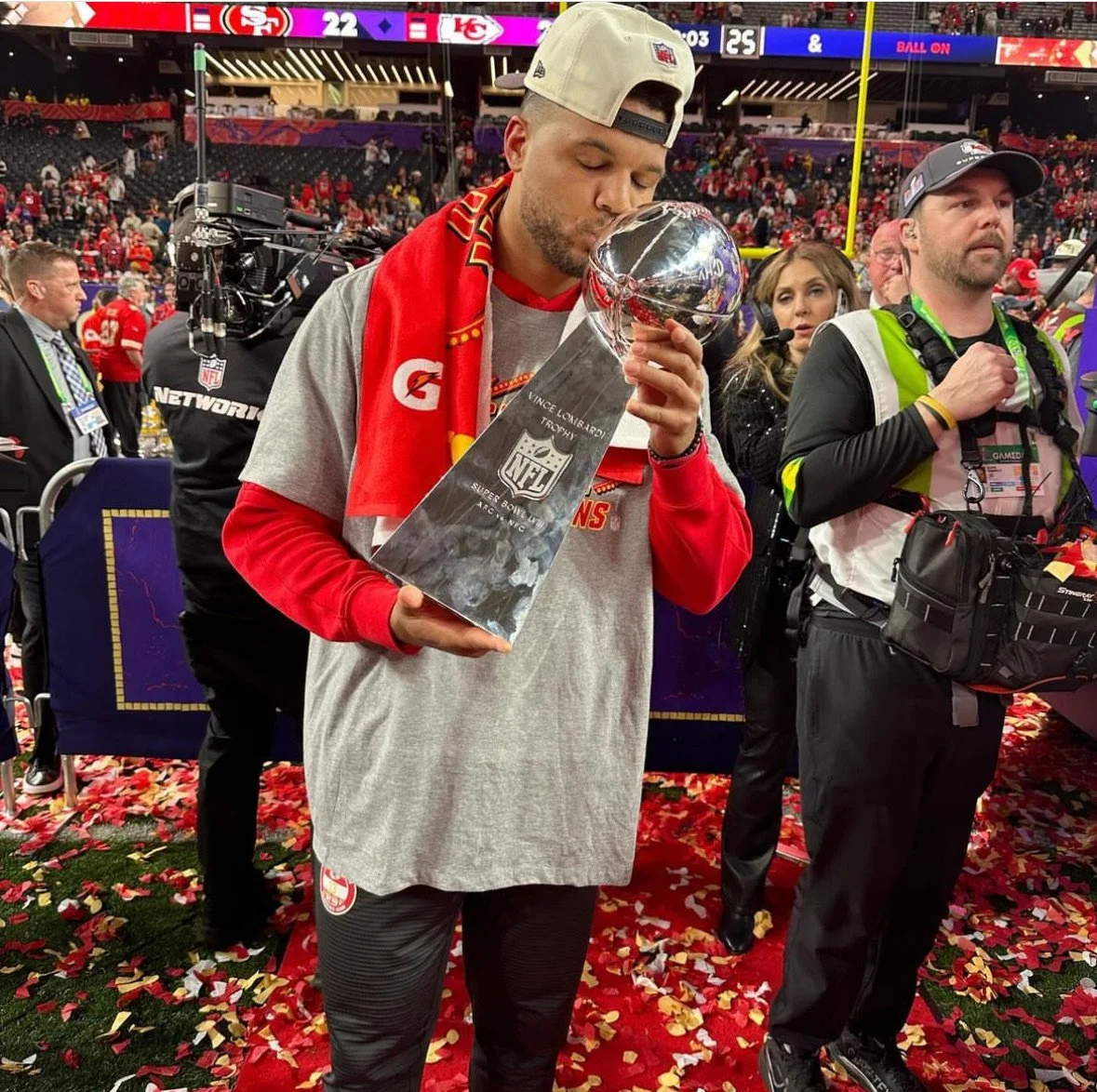 A football player kisses the Vince Lombardi Trophy at an NFL celebration event, with confetti on the ground and a crowd in the background.