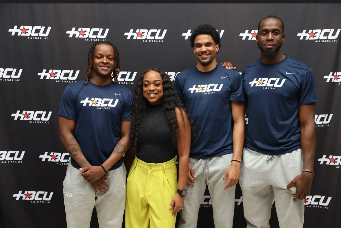 Four athletes standing in front of a black backdrop with the HBCU All Stars LLC logo. They are wearing matching navy blue T-shirts with the logo and gray sweatpants, except for one woman in yellow pants. They are smiling and posing for a group photo.