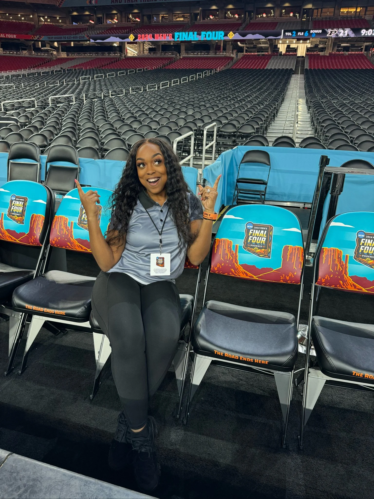 A smiling woman sitting on a chair at the 2024 NCAA men's Final Four basketball game in Phoenix, Arizona. She is wearing a gray polo shirt, black pants, and black shoes, with event badge and orange wristband. The stadium seats are mostly empty, with digital screens and signage in the background.