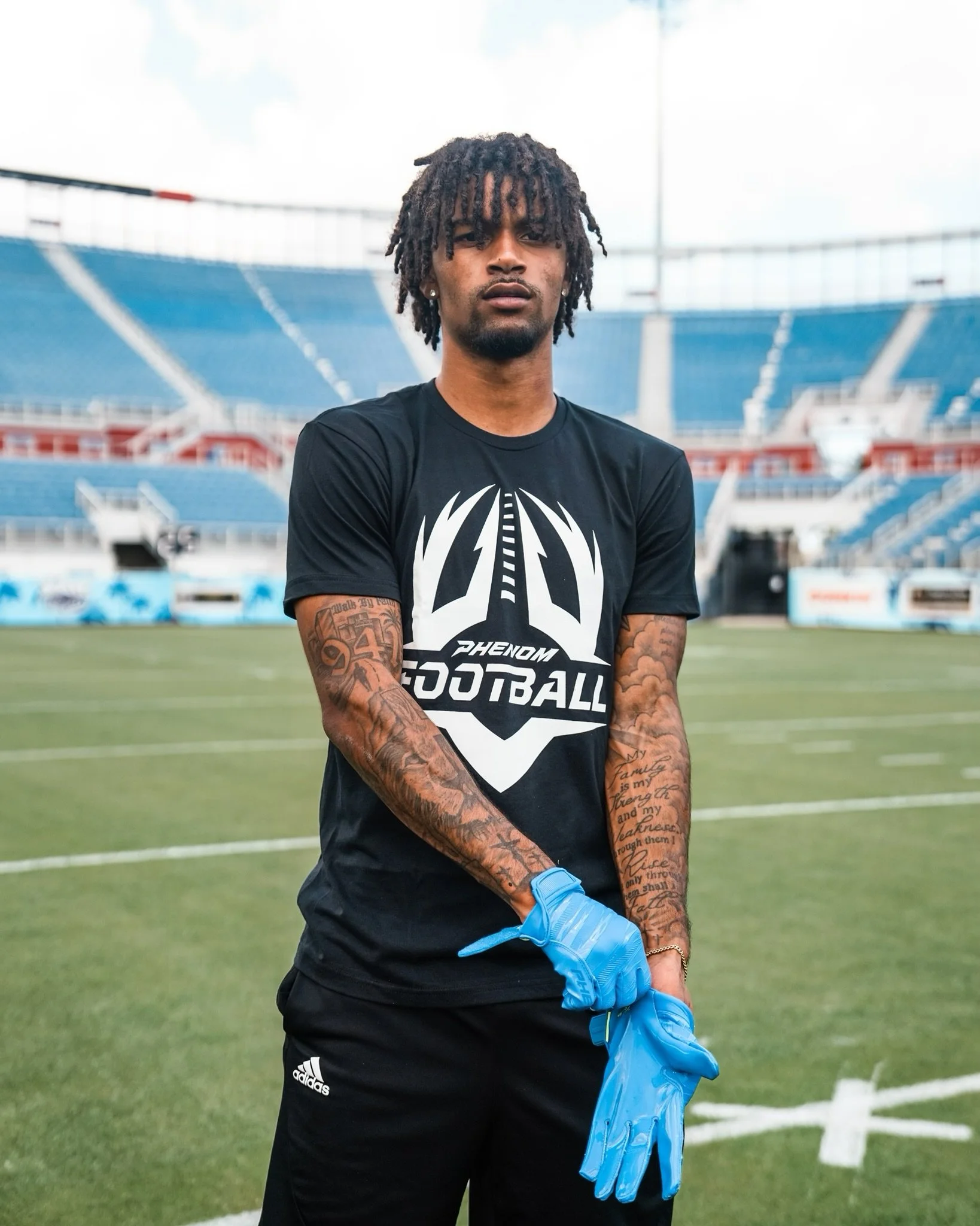 A young man with tattoos on his arms standing on a football field, wearing a black "Phenomen Football" t-shirt, black Adidas pants, and blue goalkeeper gloves.
