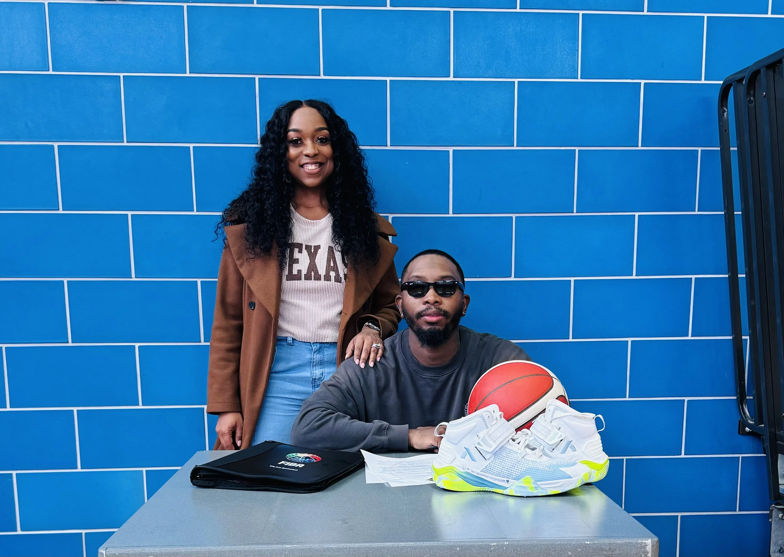 A woman standing next to a man sitting at a table with a basketball, a pair of white sneakers with neon accents, and a folder, in front of a blue tiled wall.