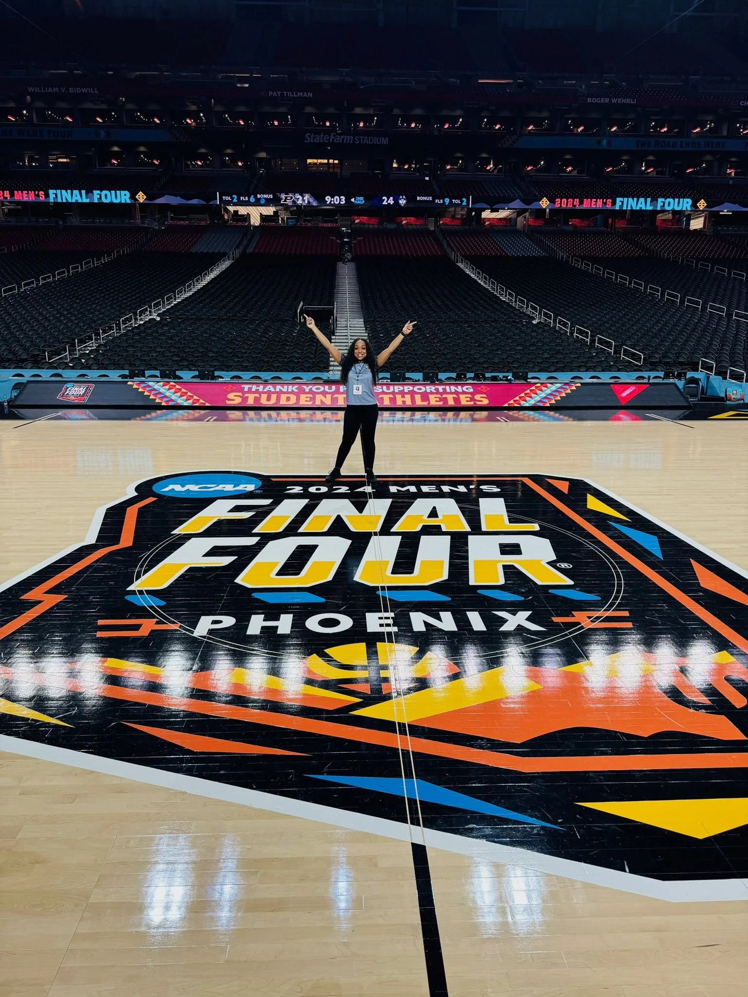 A woman standing on a basketball court with her arms raised, celebrating, at the NCAA 2024 Men's Final Four in Phoenix.