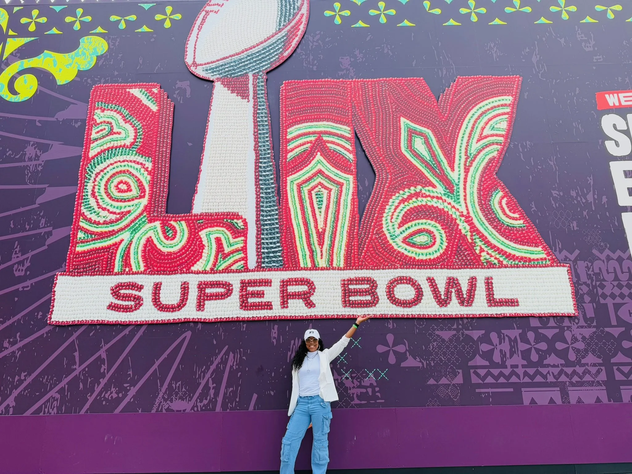 A woman with sunglasses, white blazer, white shirt, and blue pants standing in front of a large Super Bowl LVI sign on a purple background, pointing at the sign.