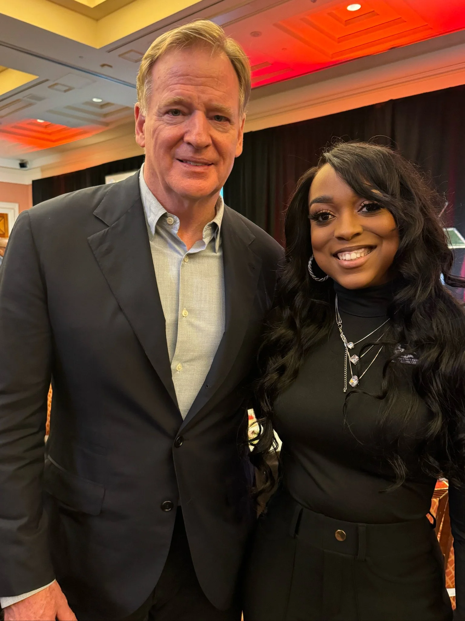 A man and woman posing together indoors, smiling for the camera at an event, with colorful lighting and a stage in the background.