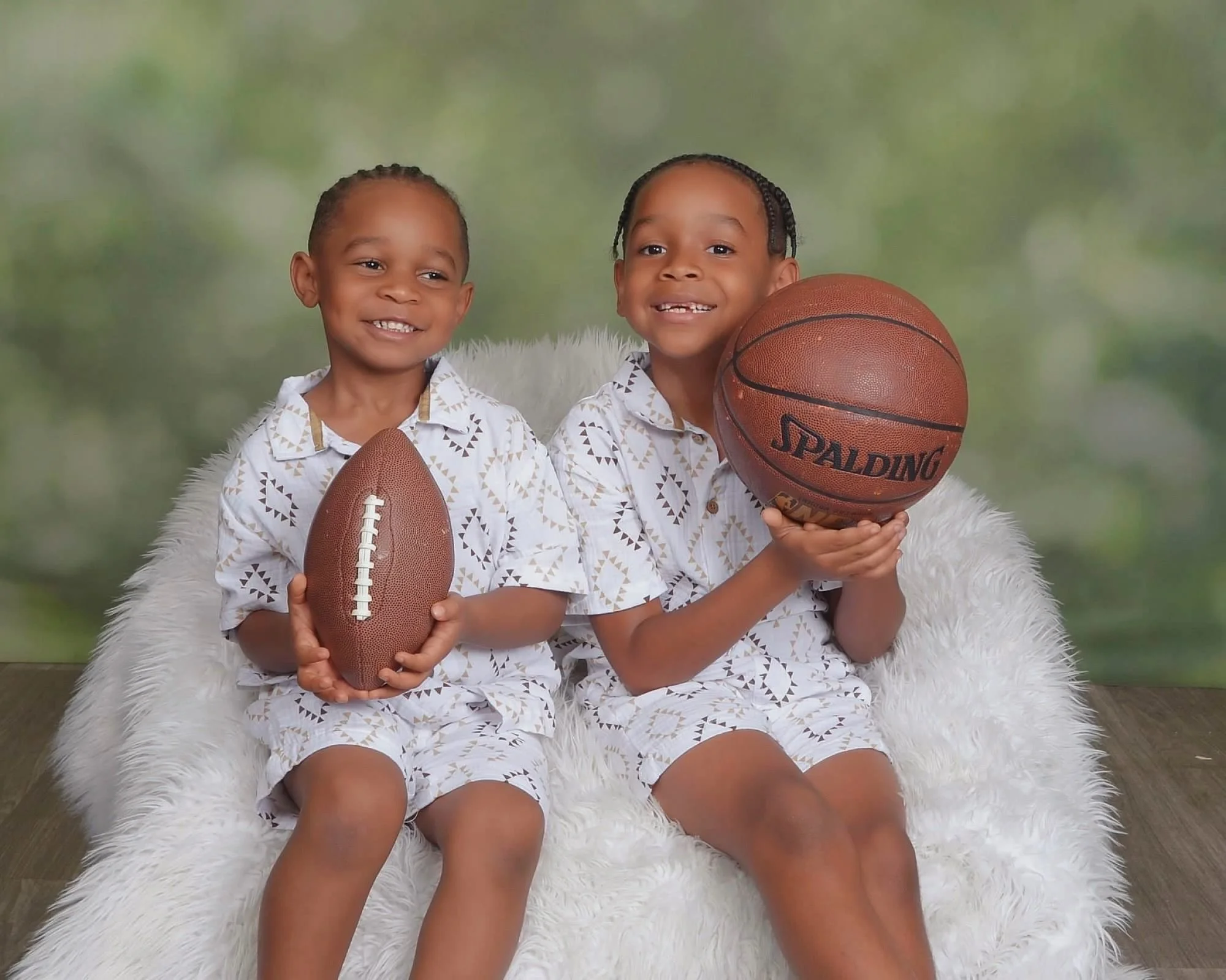 Two young children, a boy and a girl, sitting on a fluffy white chair, holding sports balls, in front of a blurred outdoor background, wearing matching patterned pajamas, smiling at the camera.