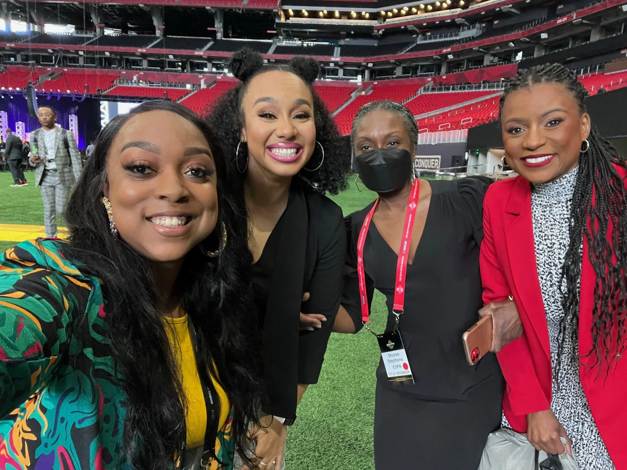 Four women standing on a football field in a stadium, smiling for a group selfie. The stadium is mostly empty with red seats and a stage in the background.