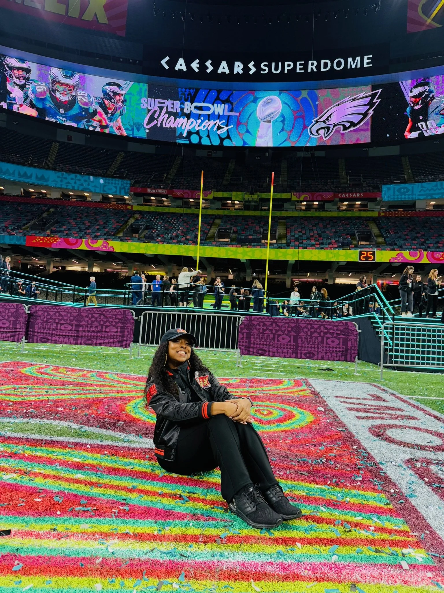 A woman sitting on a colorful, rainbow-hued area rug inside a stadium, with the Super Bowl LVII scoreboard and digital screens displaying football players and the Super Bowl Champions logo in the background.