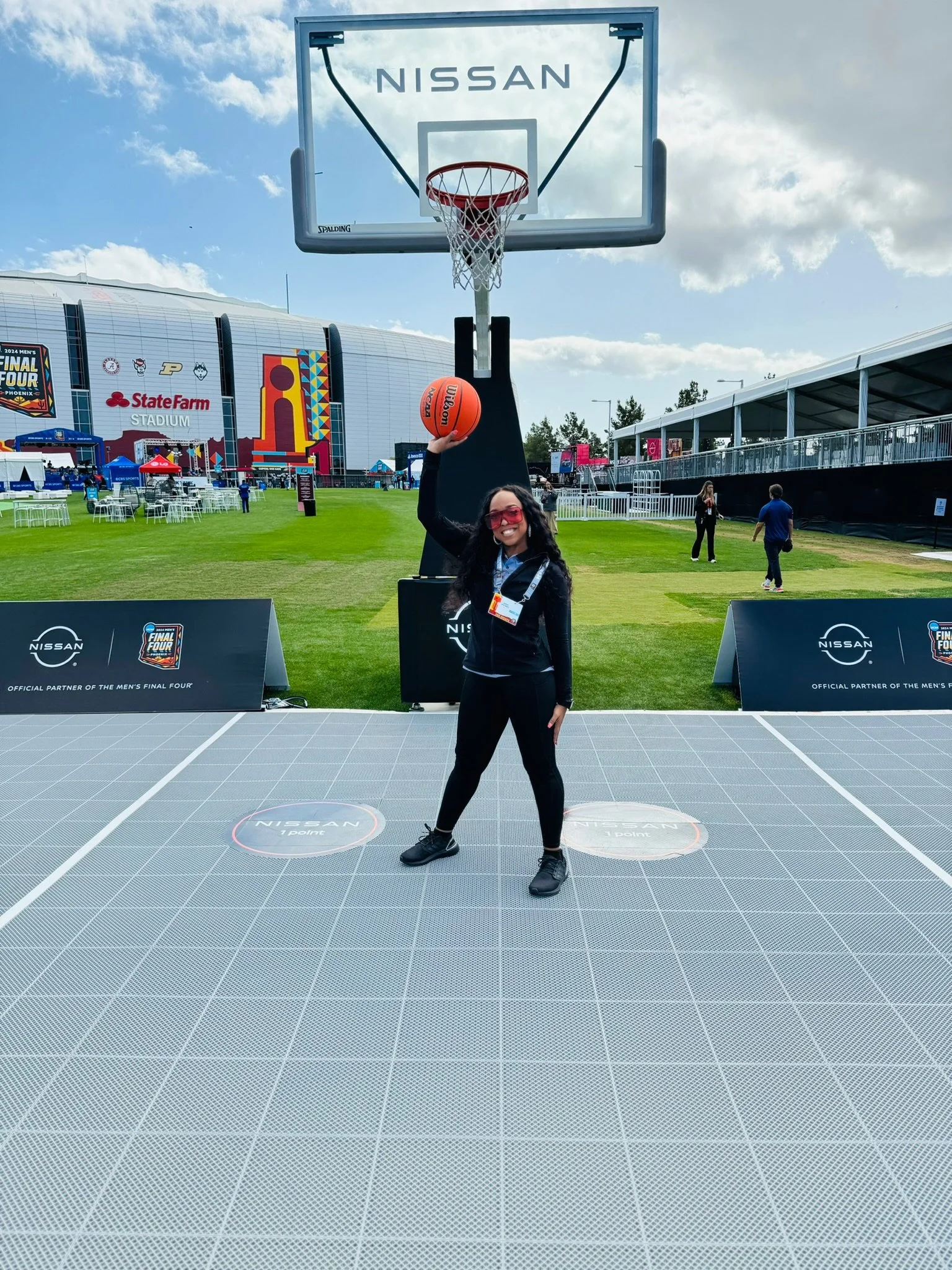Woman in black sportswear with sunglasses holding a basketball above her head in front of a basketball hoop at the Nissan-sponsored sports event outside on a sunny day.