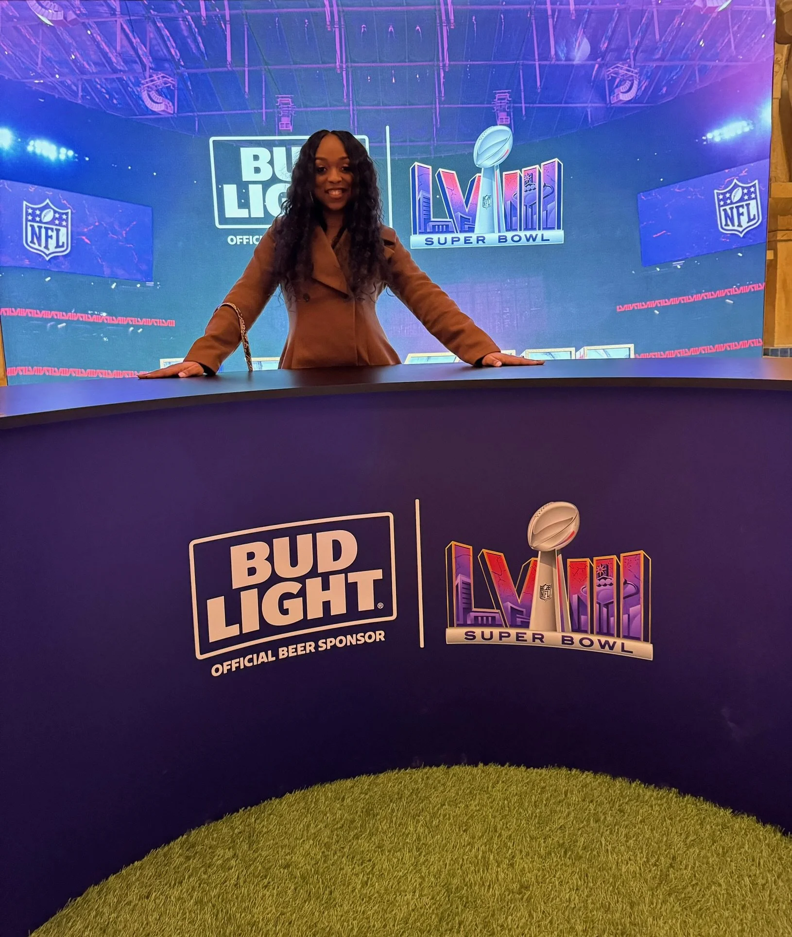A woman with long, curly black hair smiling and leaning on a table at a Super Bowl event. Behind her is a large digital display with NFL, Bud Light, and Super Bowl logos.
