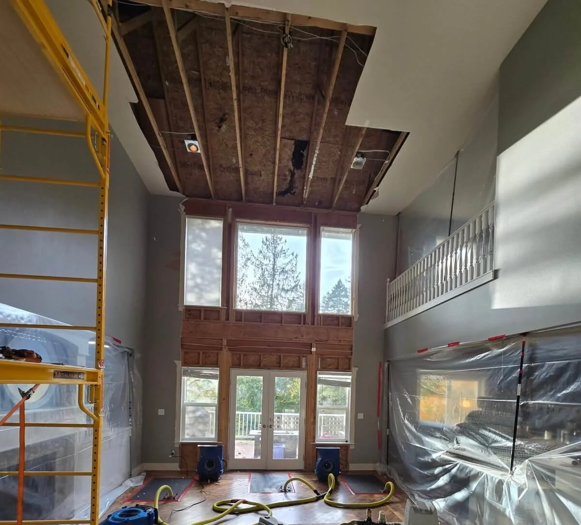 Duvall two-story room before drywall restoration, showing exposed ceiling framing, scaffolding, tall windows, and protective coverings.