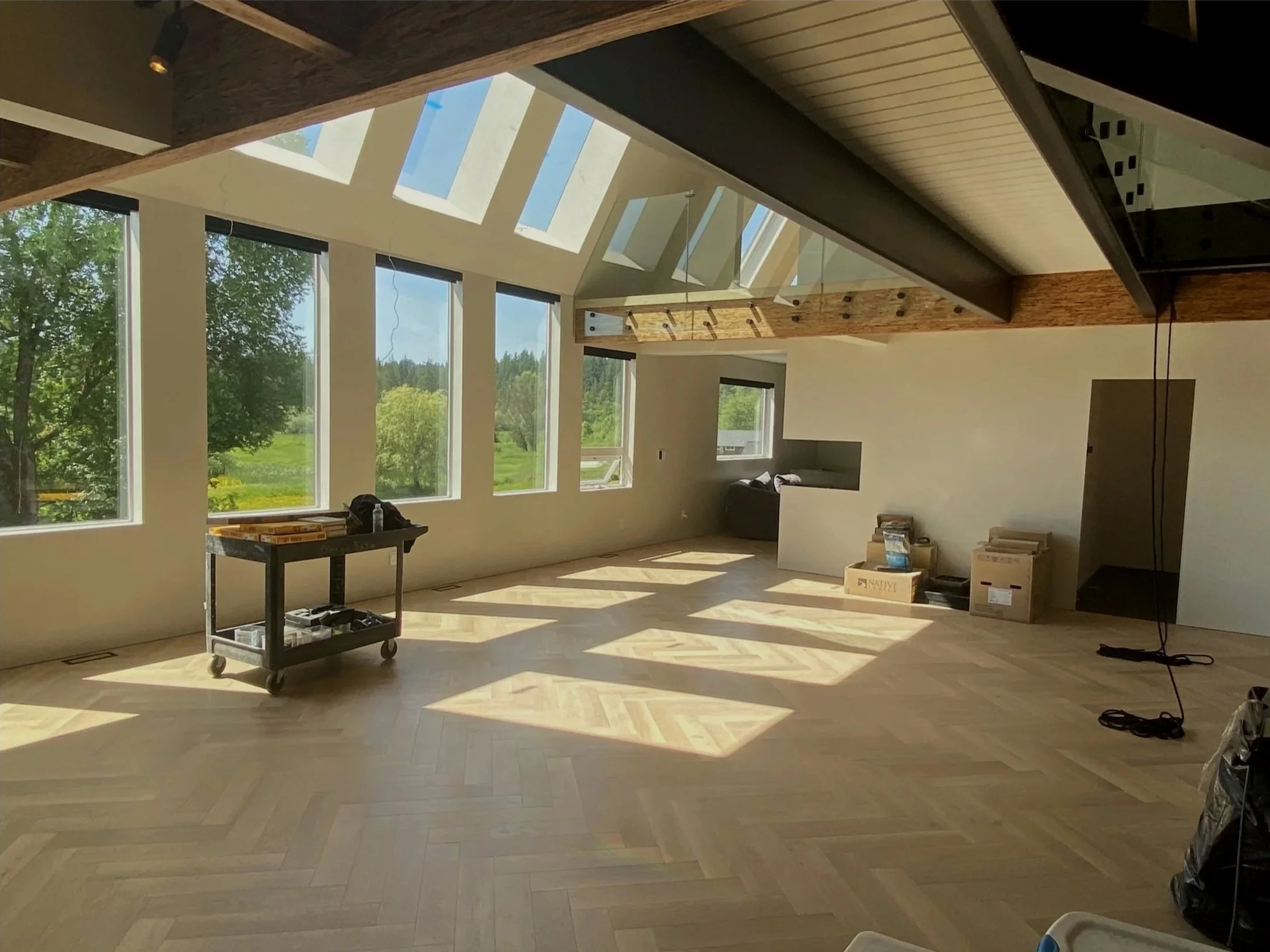 Bright, spacious room with large windows, herringbone wood floor, exposed wooden beams, and skylights. Some construction supplies and furniture boxes are scattered around.