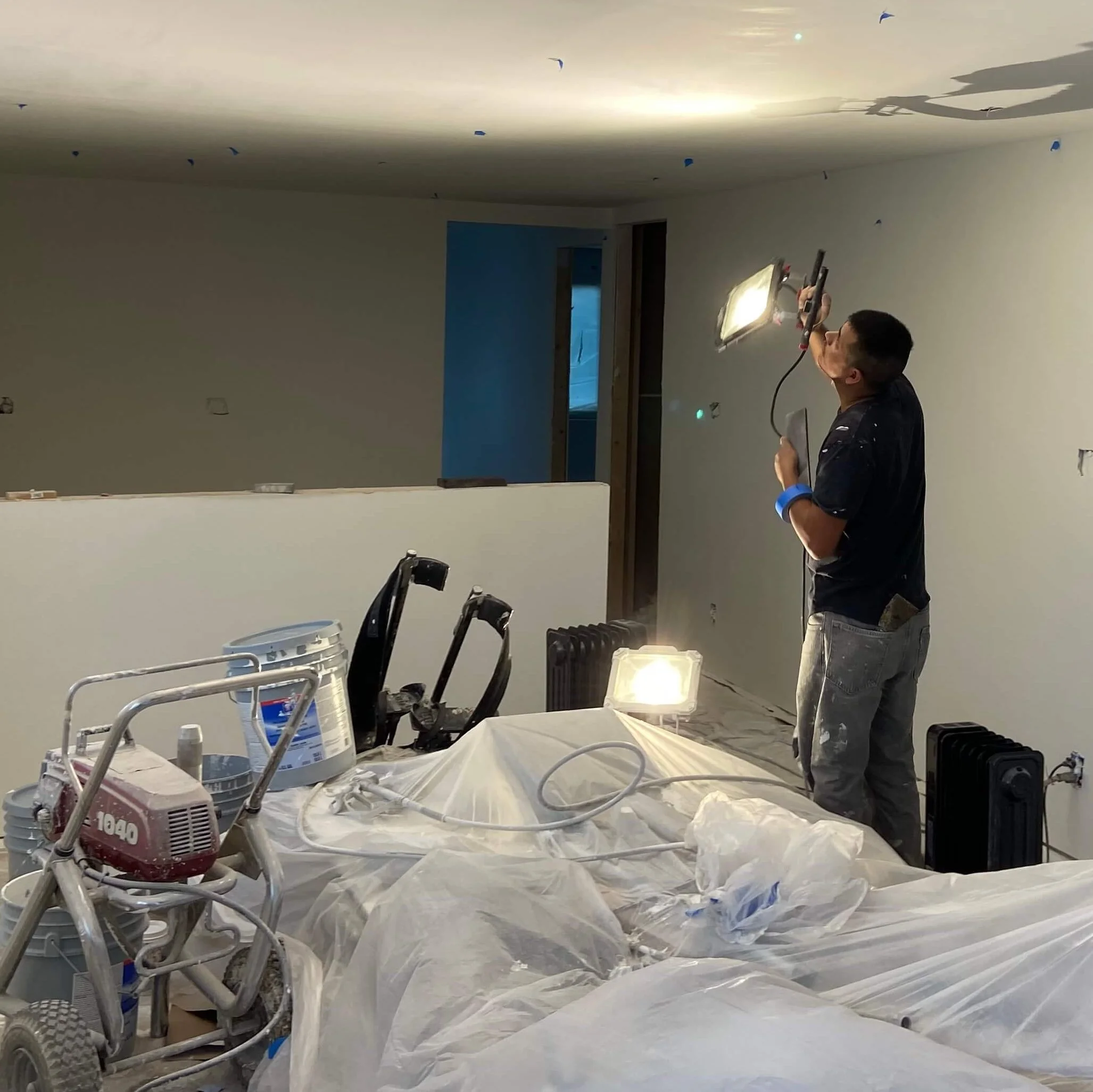 Drywall finisher inspecting walls and ceilings with a work light, with blue tape marking touch-up areas before another coat of primer.