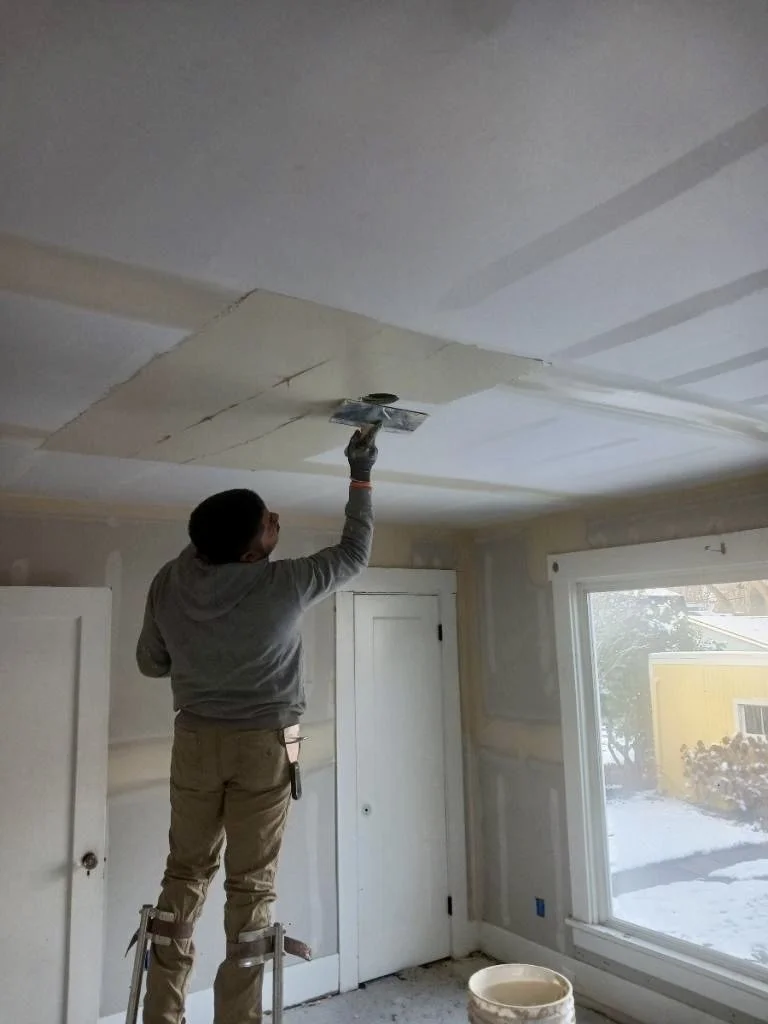 Drywall finisher applying joint compound across a ceiling with a hand trowel during full-surface skim coating.
