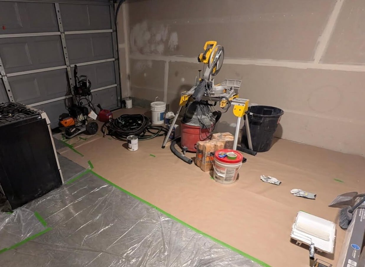 Seattle garage with visible builder-grade drywall joints, tools, and protective floor coverings before refinishing.