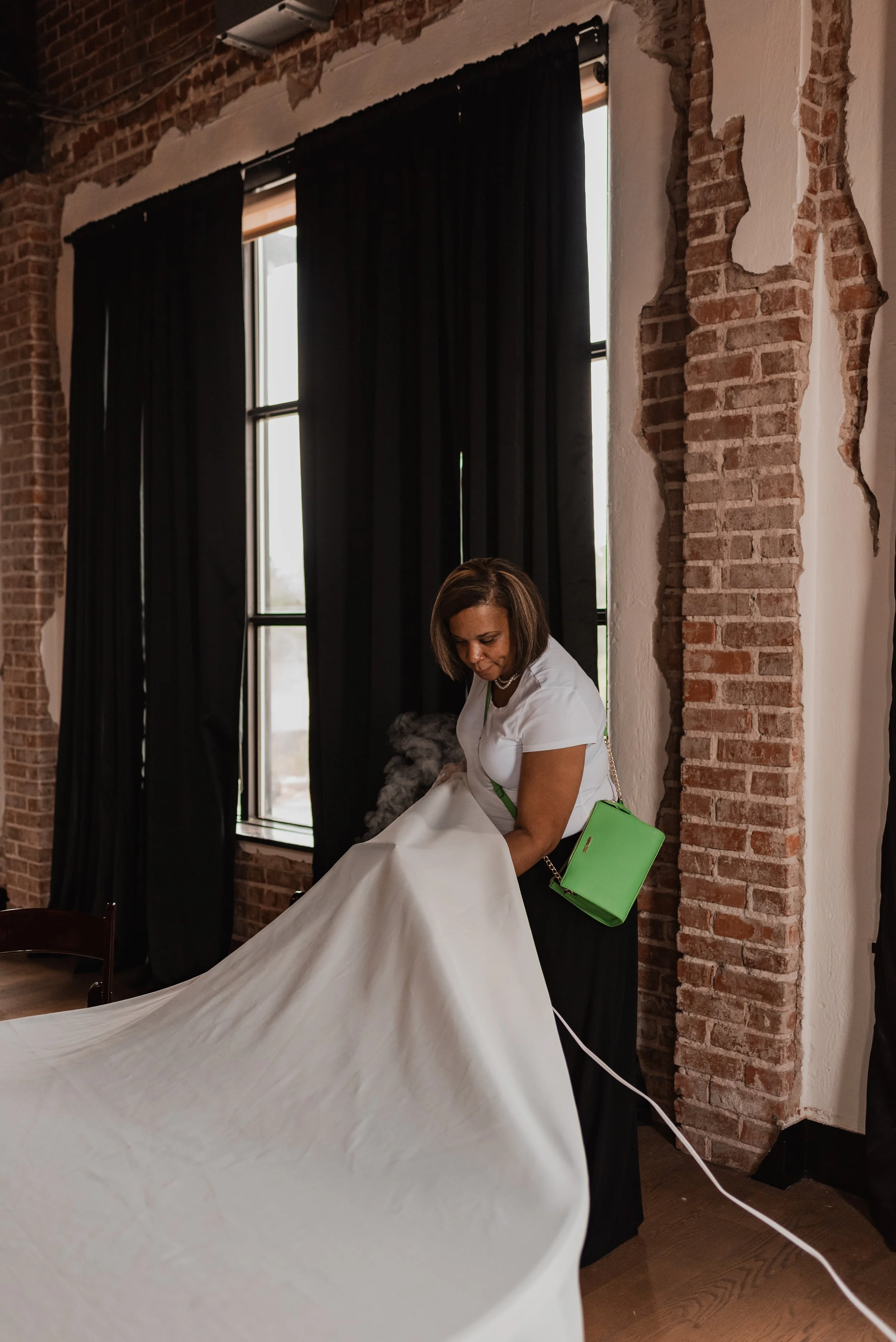 A woman with shoulder-length brown hair, wearing a white shirt, is steaming a white wedding dress indoors near large windows with black curtains. The room has exposed brick walls and a rustic aesthetic.