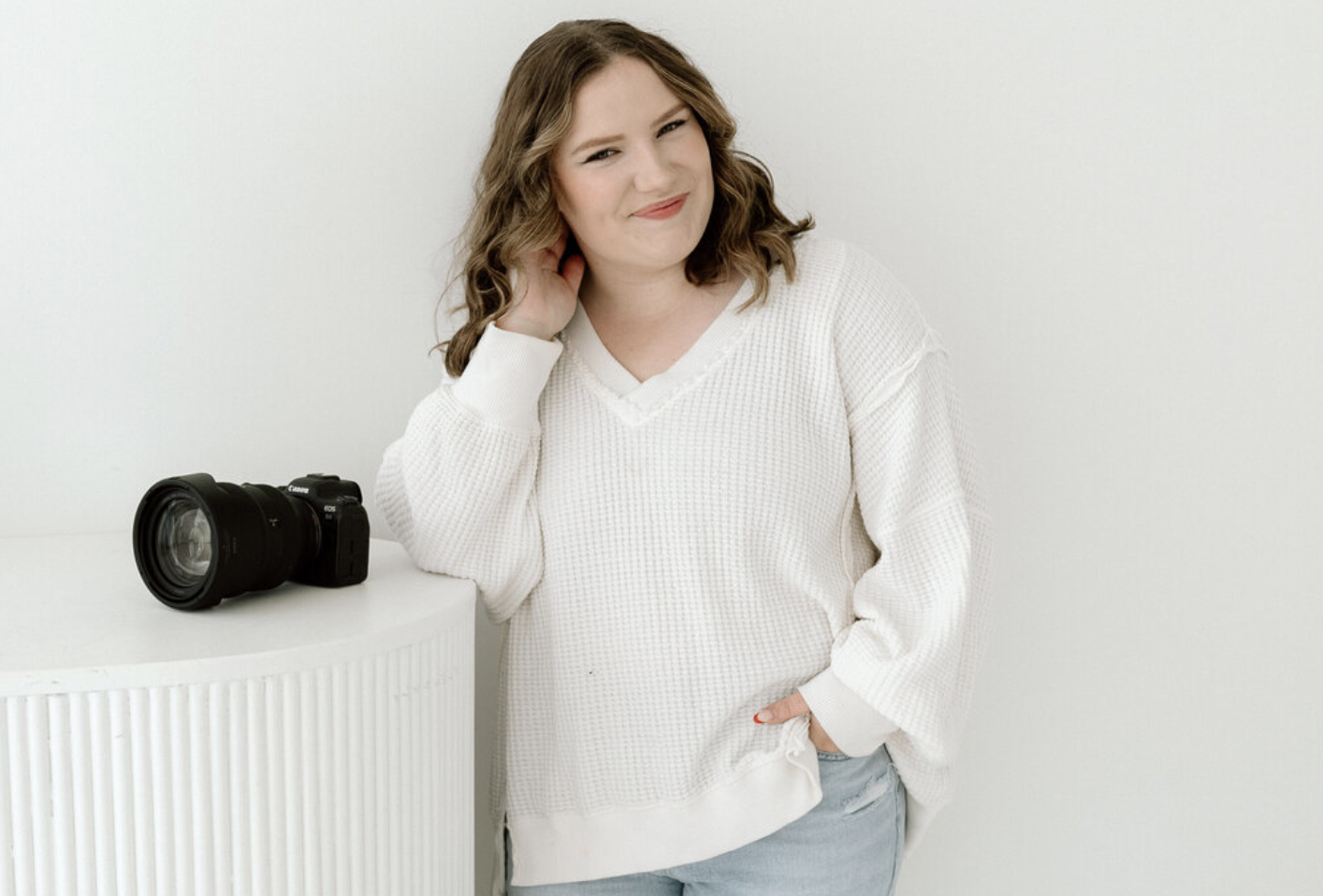 A young woman with wavy light brown hair, wearing a white knitted sweater and light jeans, smiling and standing against a white background. A professional camera is placed on a white surface next to her.