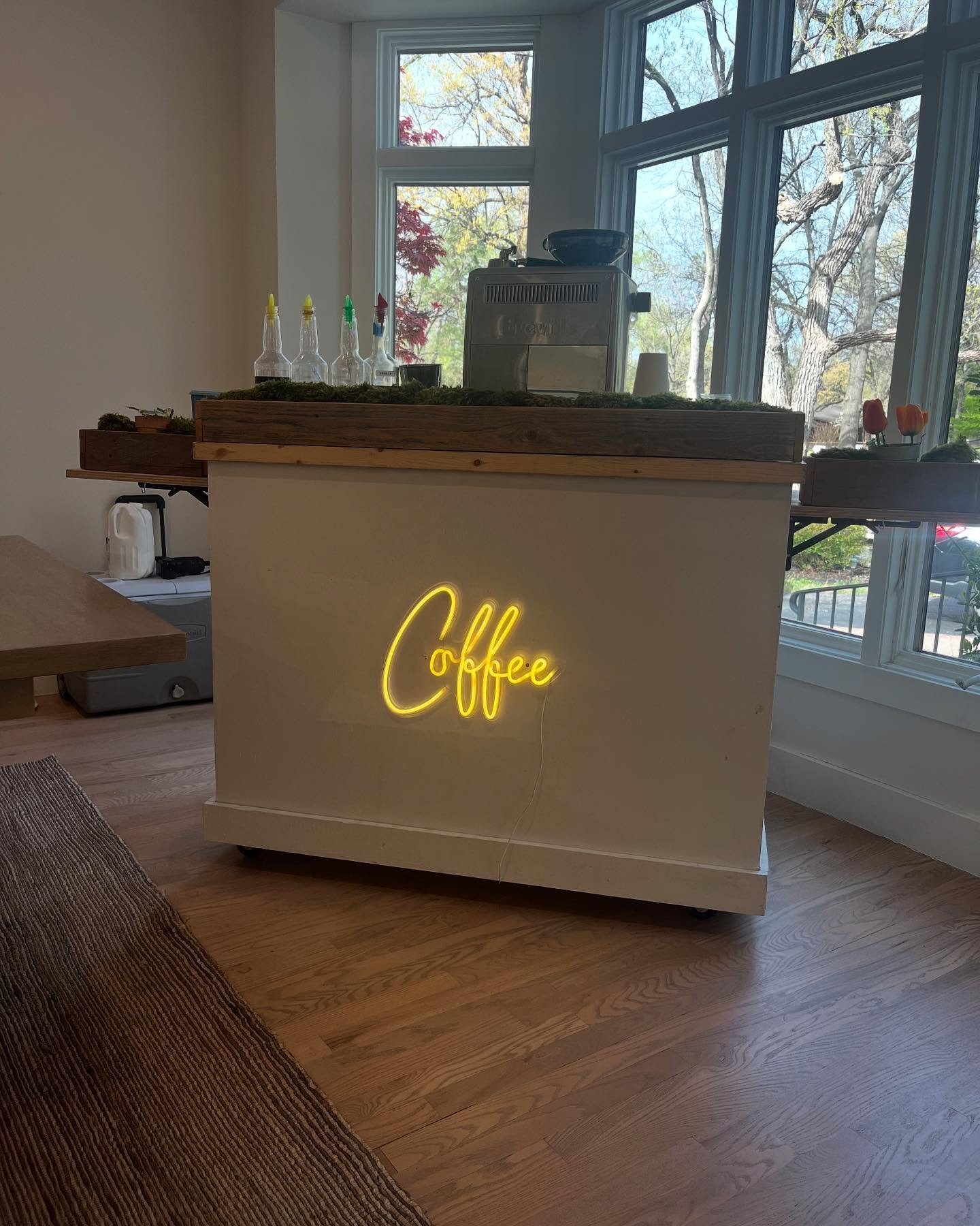 A cafe counter with a yellow neon sign that says 'Coffee,' situated in front of large windows showing an outdoor view with trees and blue sky.