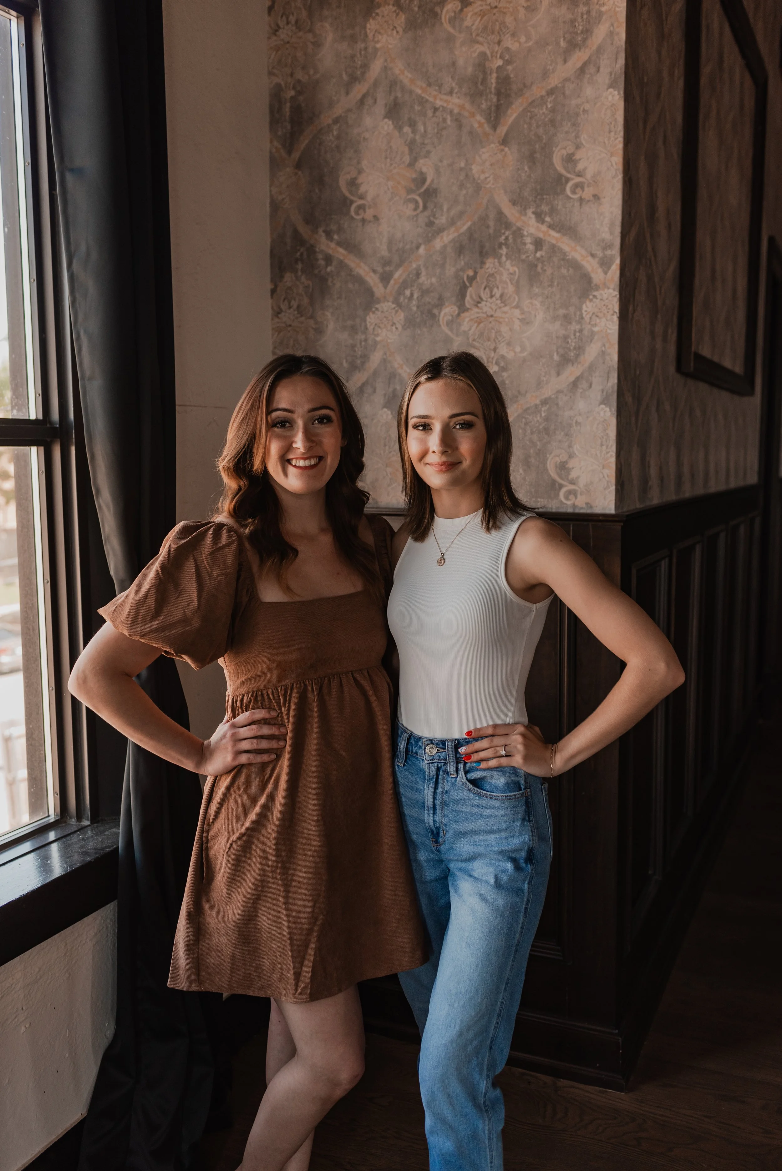 Two young women standing indoors near a window, smiling at the camera. One wears a brown dress with puffed sleeves, and the other wears a white sleeveless top with blue jeans.