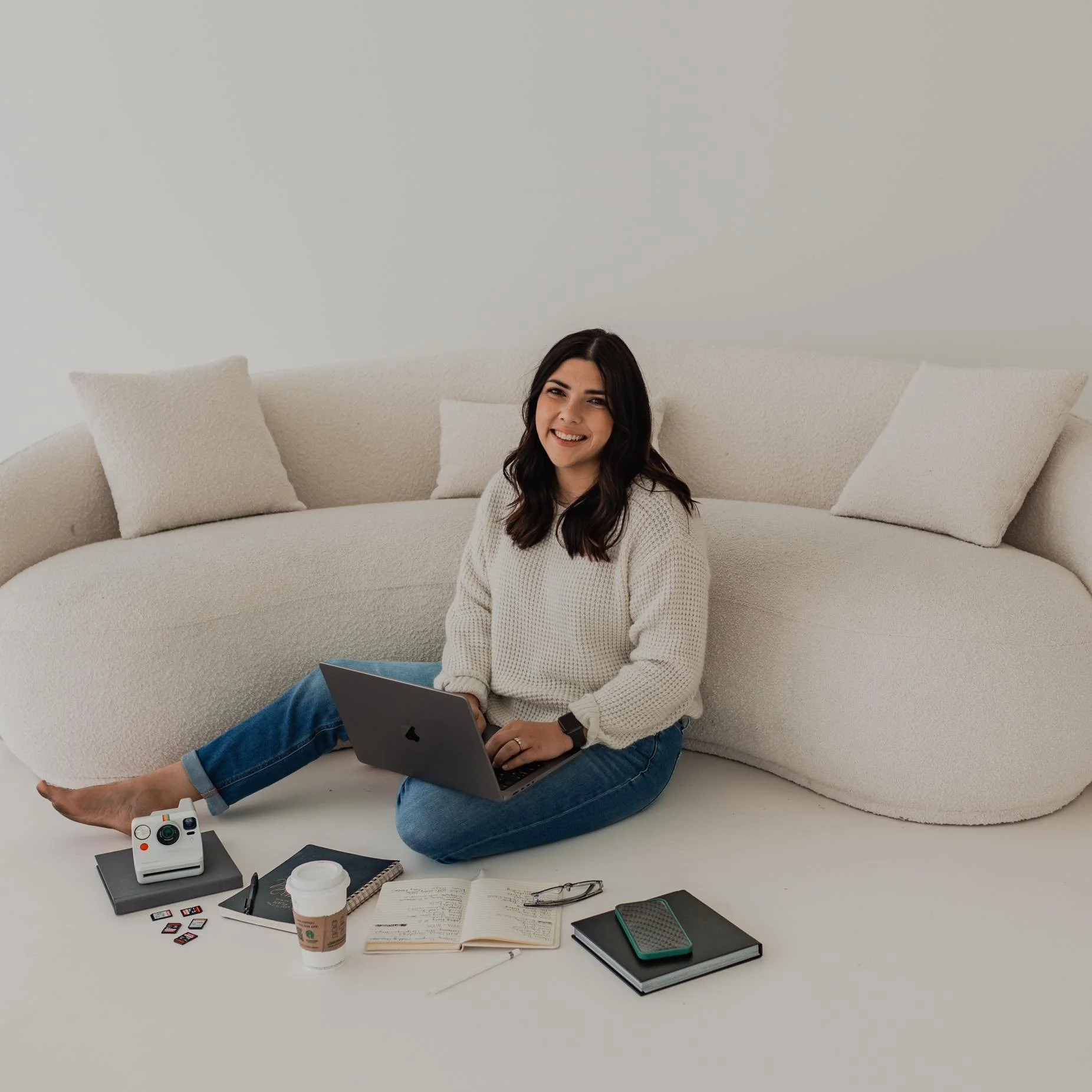 A smiling woman sitting cross-legged on the floor in front of a cream-colored sofa, surrounded by a laptop, camera, notebooks, a coffee cup, glasses, and a smartphone.