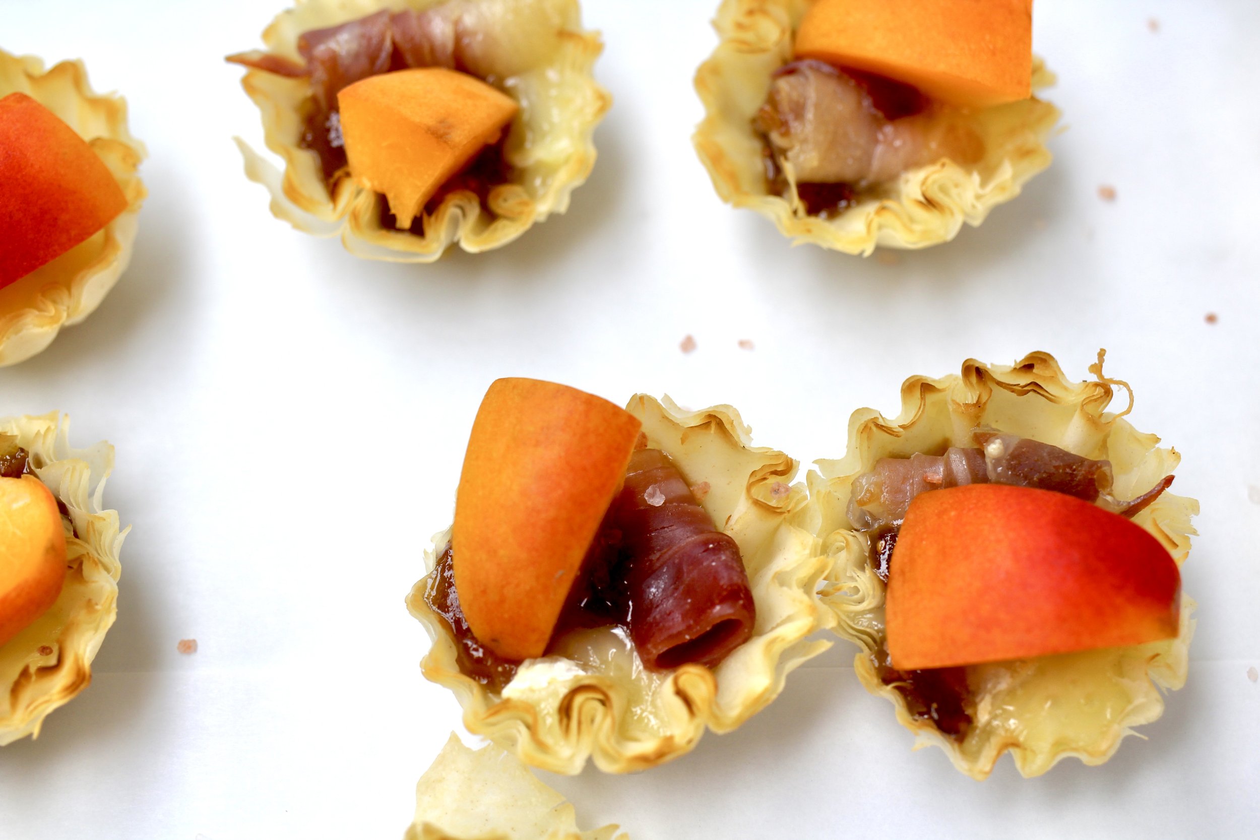 Mini tartlets filled with caramelized onions, topped with slices of peach and prosciutto, arranged on a white surface.