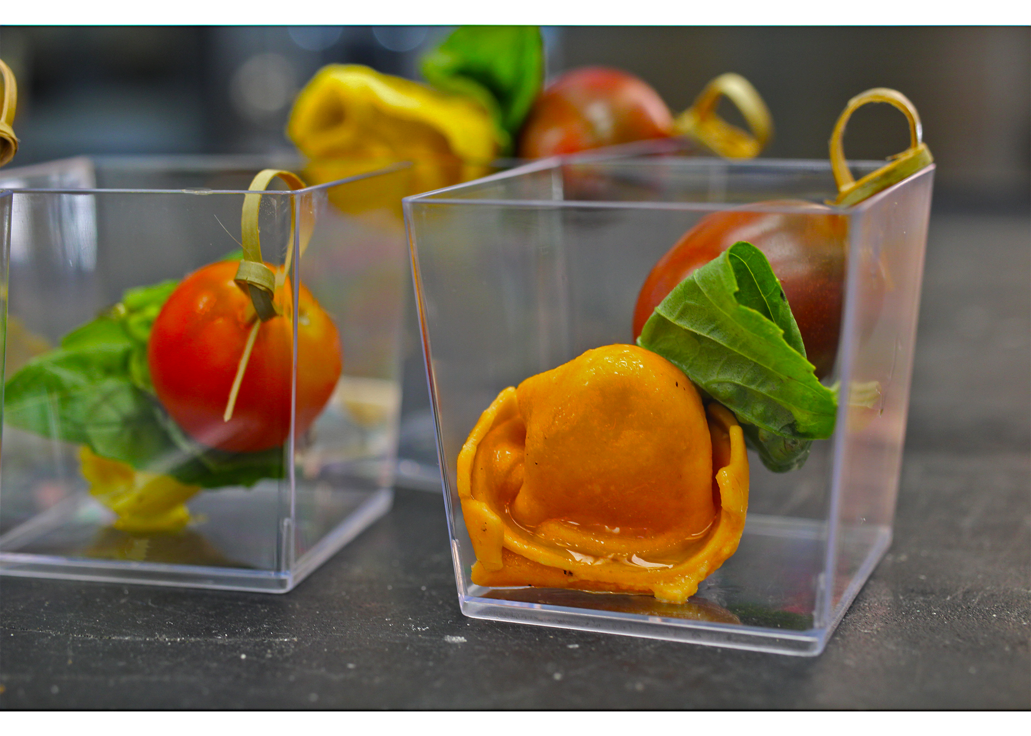 Close-up of plastic containers holding preserved cherry tomatoes wrapped with yellow spinach leaves, placed on a dark surface.
