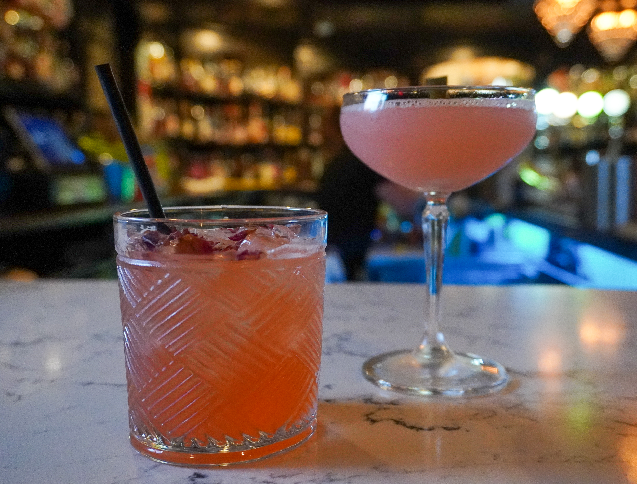 Two cocktails at The Bellview with a blurred bar background, one in a short glass with ice and a straw, and the other in a coupe glass.