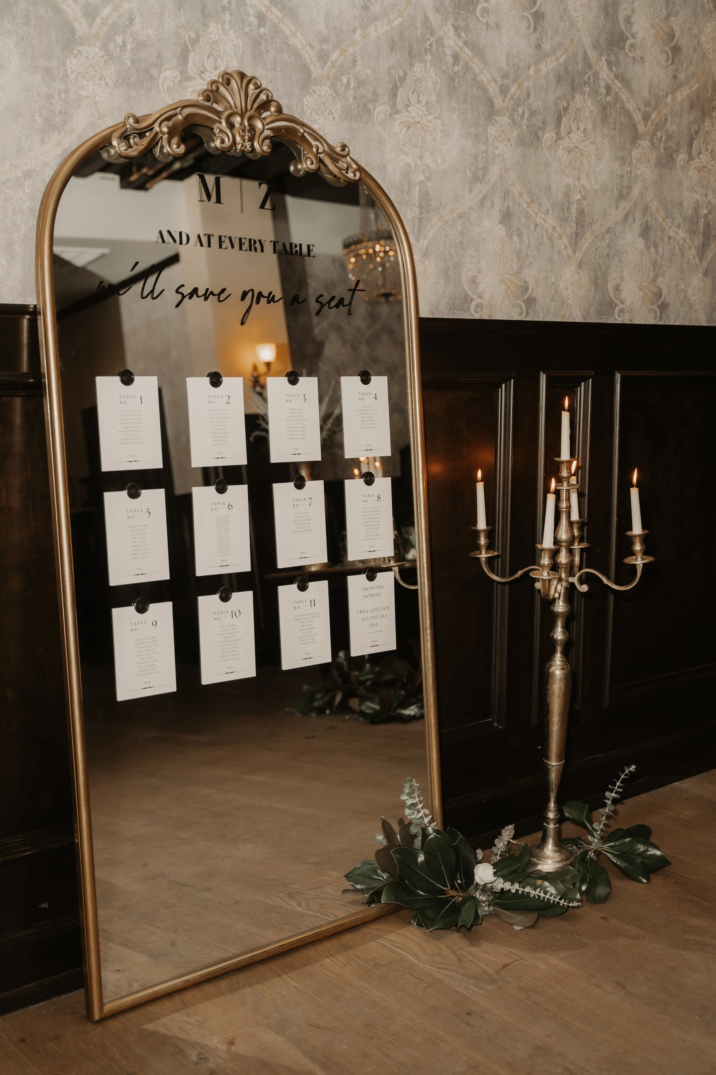 Decorative standing mirror reflecting a seating chart and a candelabrum with lit candles, placed on a wooden floor near dark wooden paneling and ornate wallpaper.