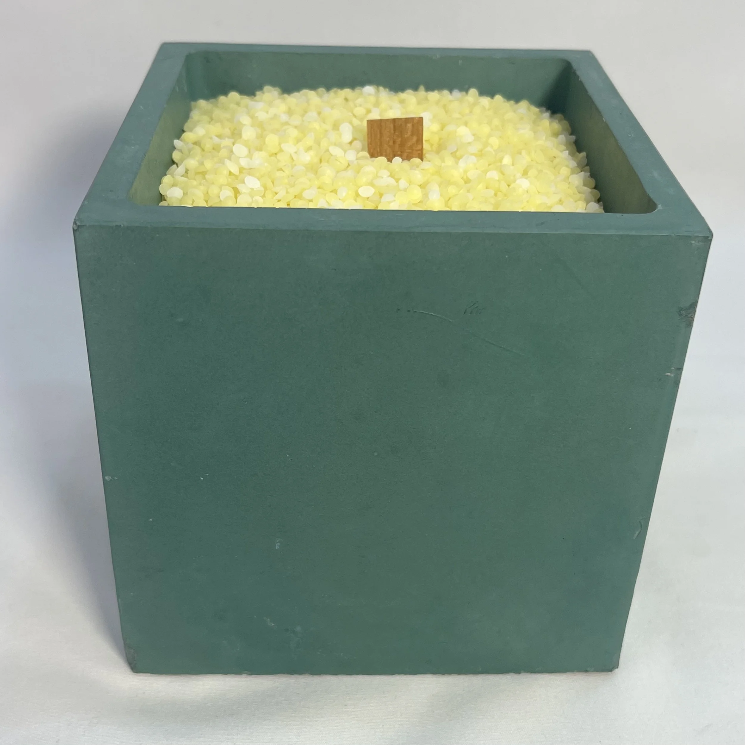green concrete cube filled with beeswax grains and one wooden wick in the middle of the grains