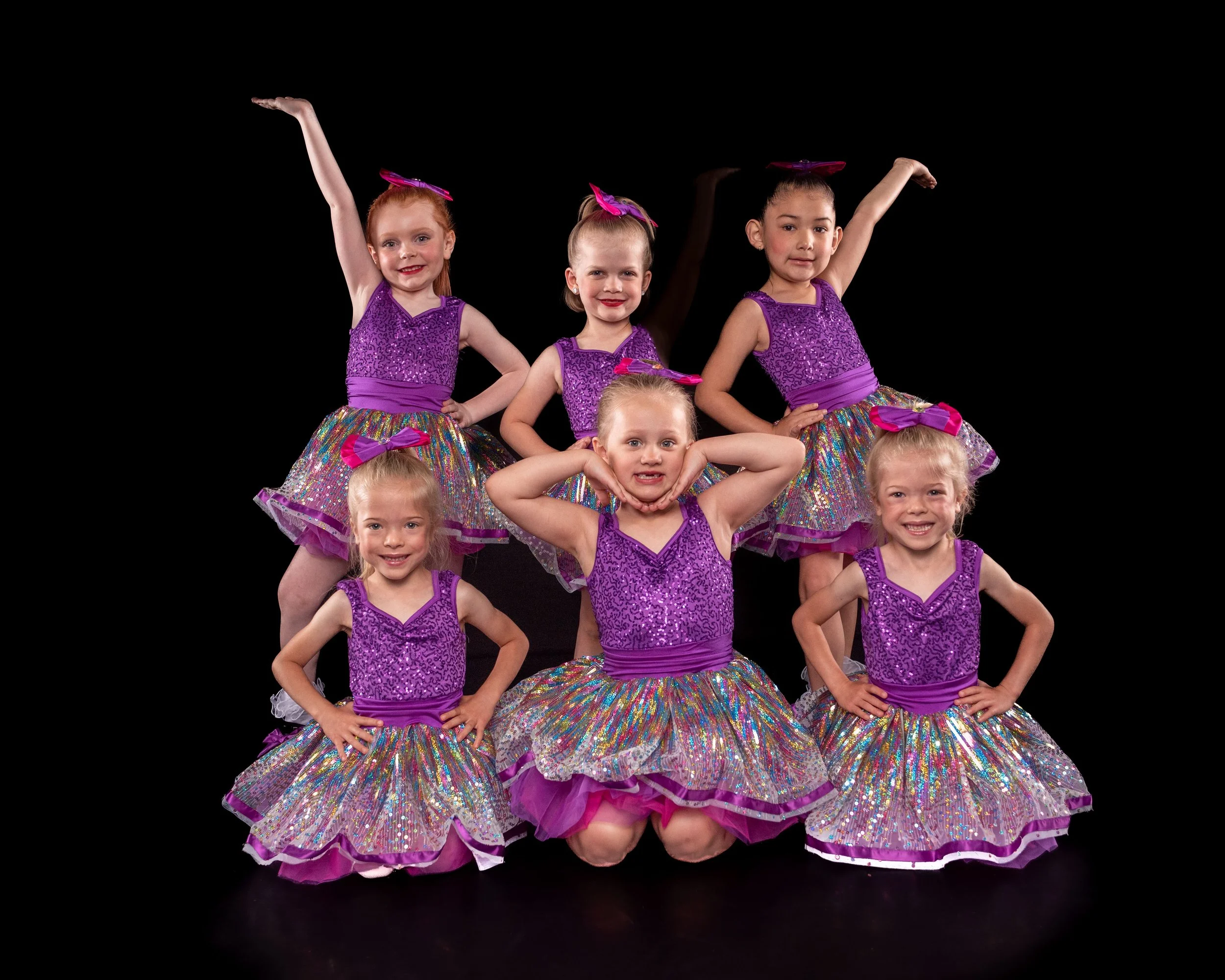 Group of young girls in colorful, sequined ballet costumes with pink bows, posing against a black background.