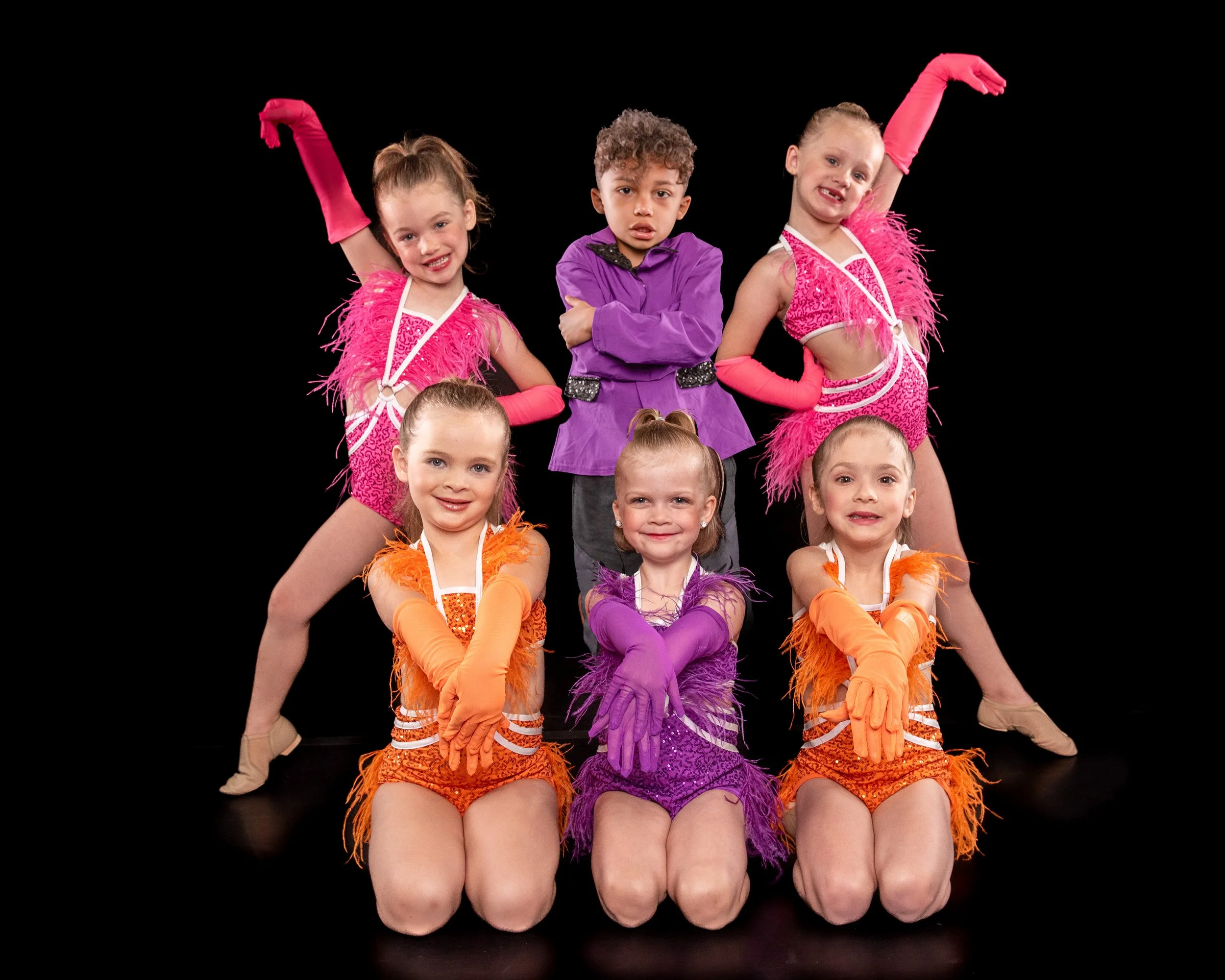 Group of young children dressed in colorful costumes, seated and standing on a black background, smiling and posing for the camera.