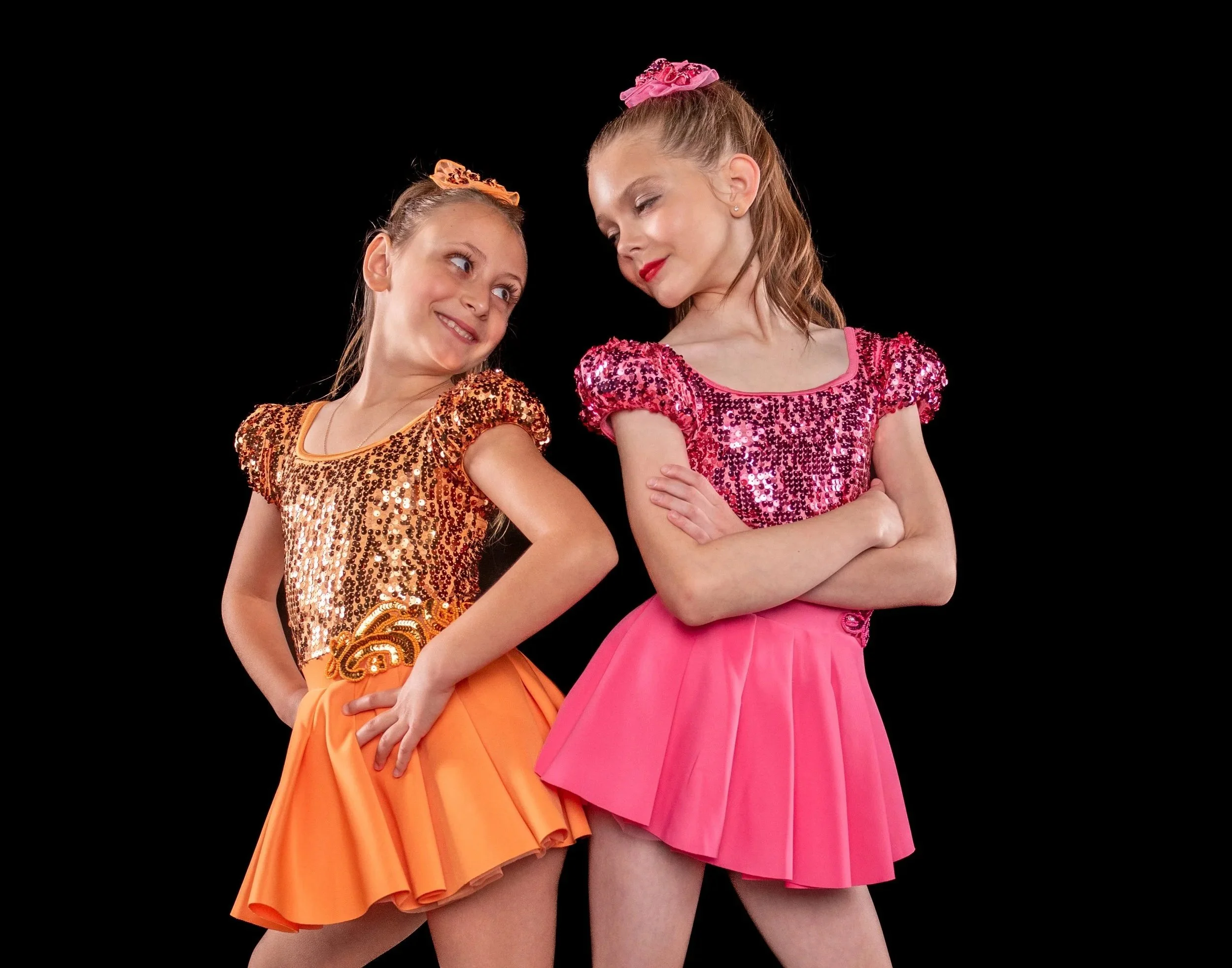 Two young girls dressed in colorful, sequin-embellished dresses pose playfully against a black background.