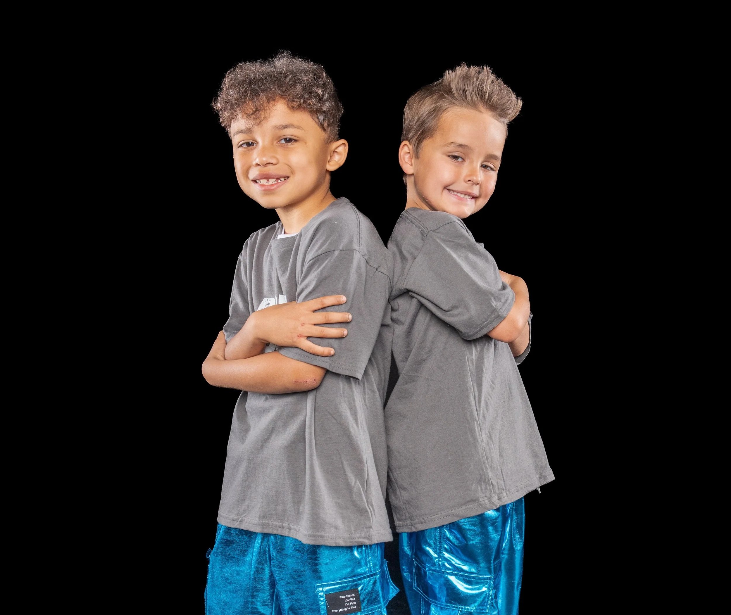 Two young boys stand back to back with arms crossed, smiling on a black background.